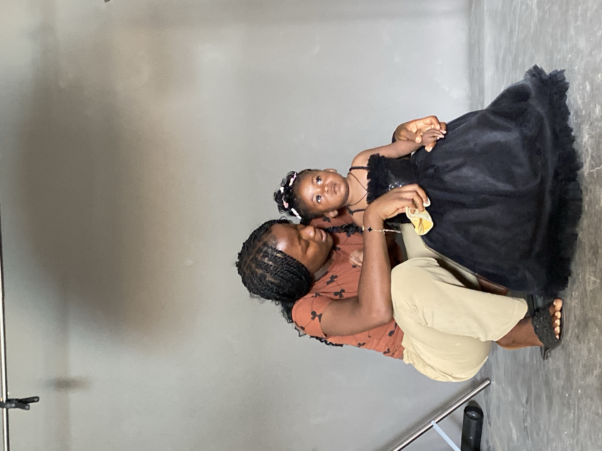 A woman sits on the floor in a gray room, holding a toddler dressed in a black dress. The woman wears a brown t-shirt with black bow tie patterns and beige pants. She has braided hair and a bracelet on her left wrist. The toddler has a headband with white and pink flowers. The floor is gray concrete. There is a metal photography stand in the lower left of the frame. This image was taken in Jos, Nigeria.
