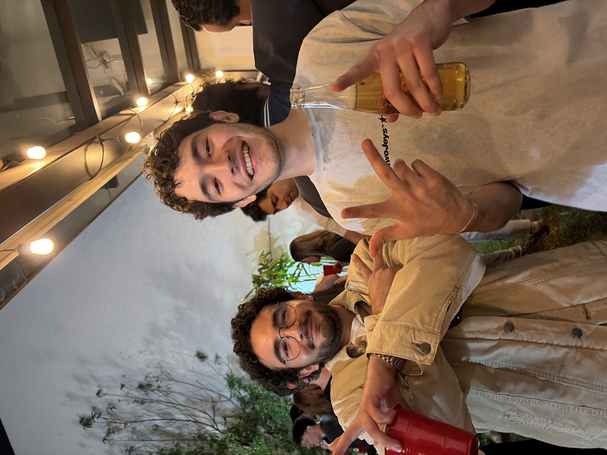 Two young men, one holding a bottle of beer, the other a red cup, are posing for a photo at an outdoor gathering. The man with the beer is wearing a light gray shirt and holding up two fingers in a peace sign. The man with the red cup is wearing glasses and a tan jacket. In the background, other people are gathered around, and there is foliage and string lights. The location appears to be a yard or patio. The city and state are unknown.
