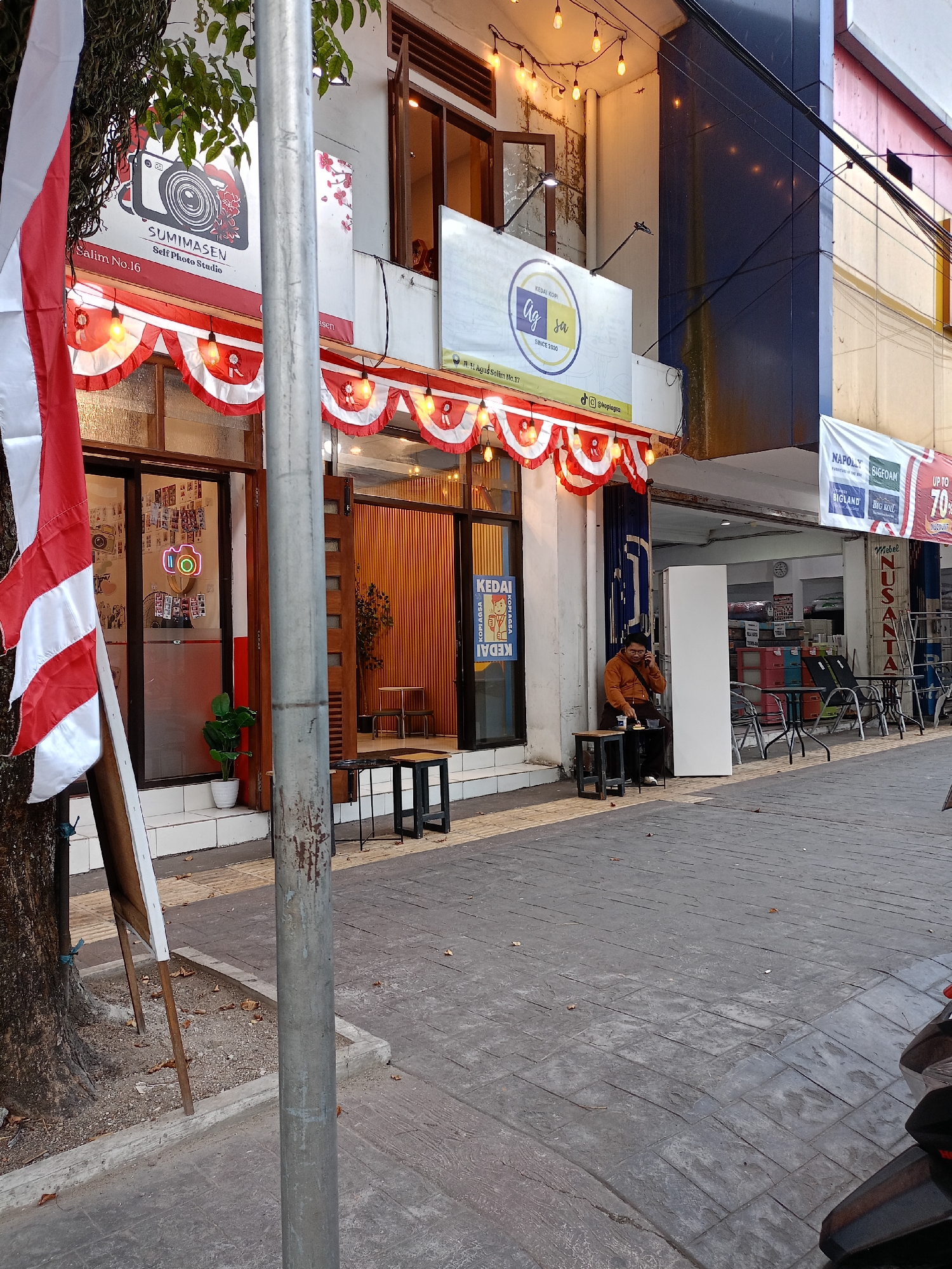 The image depicts the facade of a commercial establishment in Batu, Indonesia, with an Indonesian flag prominently displayed on the left. The building features a white exterior with red and white decorative bunting across its upper facade, strung with lights. Two businesses are visible: "SUMIMASEN Self Photo Studio" with a logo of a camera and floral motifs, and "KEDAI KOPI ag jà" with a circular logo featuring the text "ag jà SINCE 2020". A man wearing an orange jacket is seated at a small table outside the coffee shop, engaged in a phone call. The street in front of the establishments is paved with textured grey tiles. In the background, a furniture store, "Meubel NUSANTARA", is visible with various furniture items displayed outside. The scene appears to be a street-level view of a commercial area in Batu, likely during daylight hours.