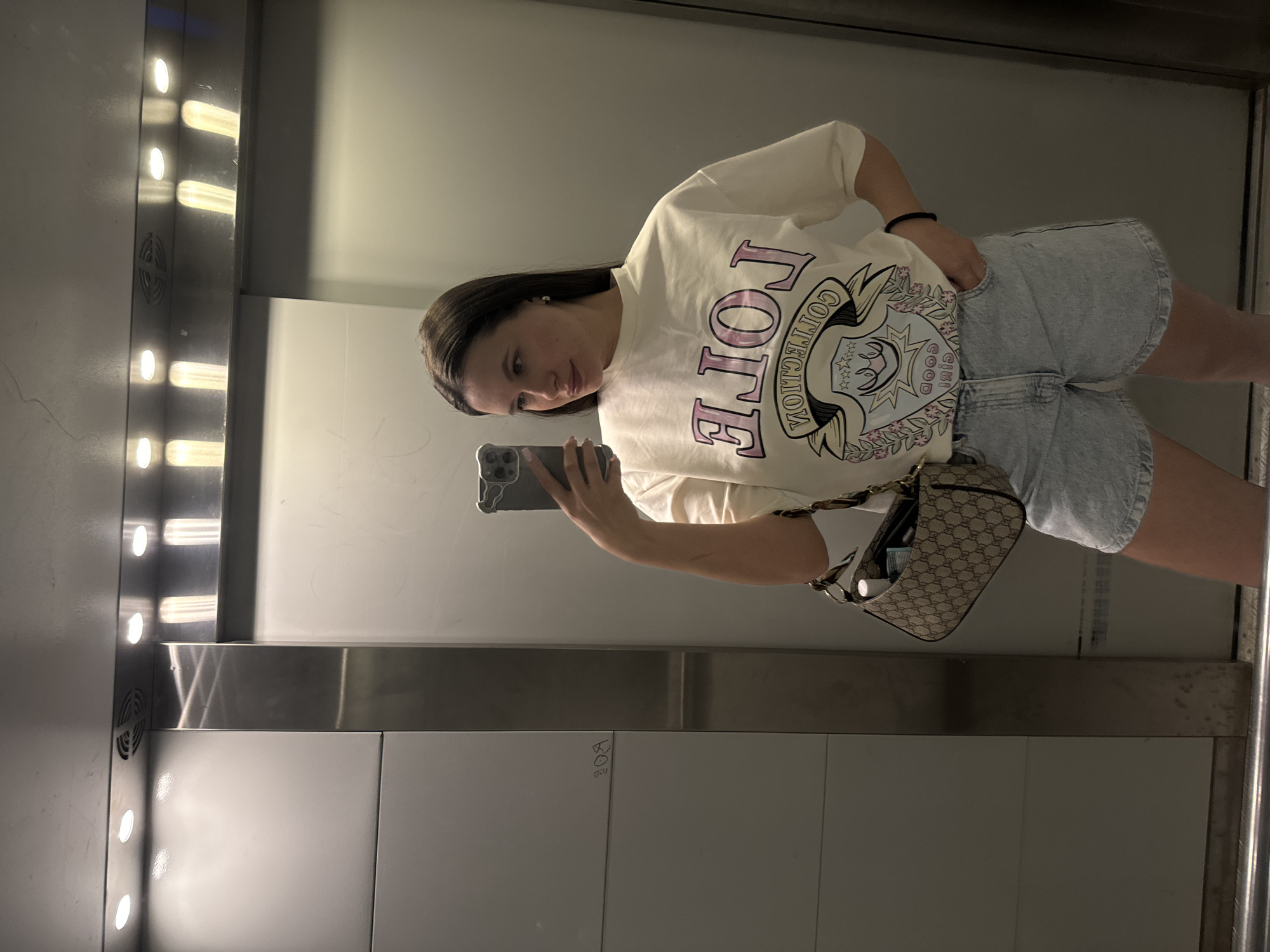 A young woman stands in an elevator, holding a smartphone to take a selfie. She is wearing a white t-shirt with a graphic design, light blue denim shorts, and a small patterned shoulder bag. The elevator has a metallic interior with vertical rows of lights. Writing is visible on the wall behind her. The location is Odintsovo, Russia.
