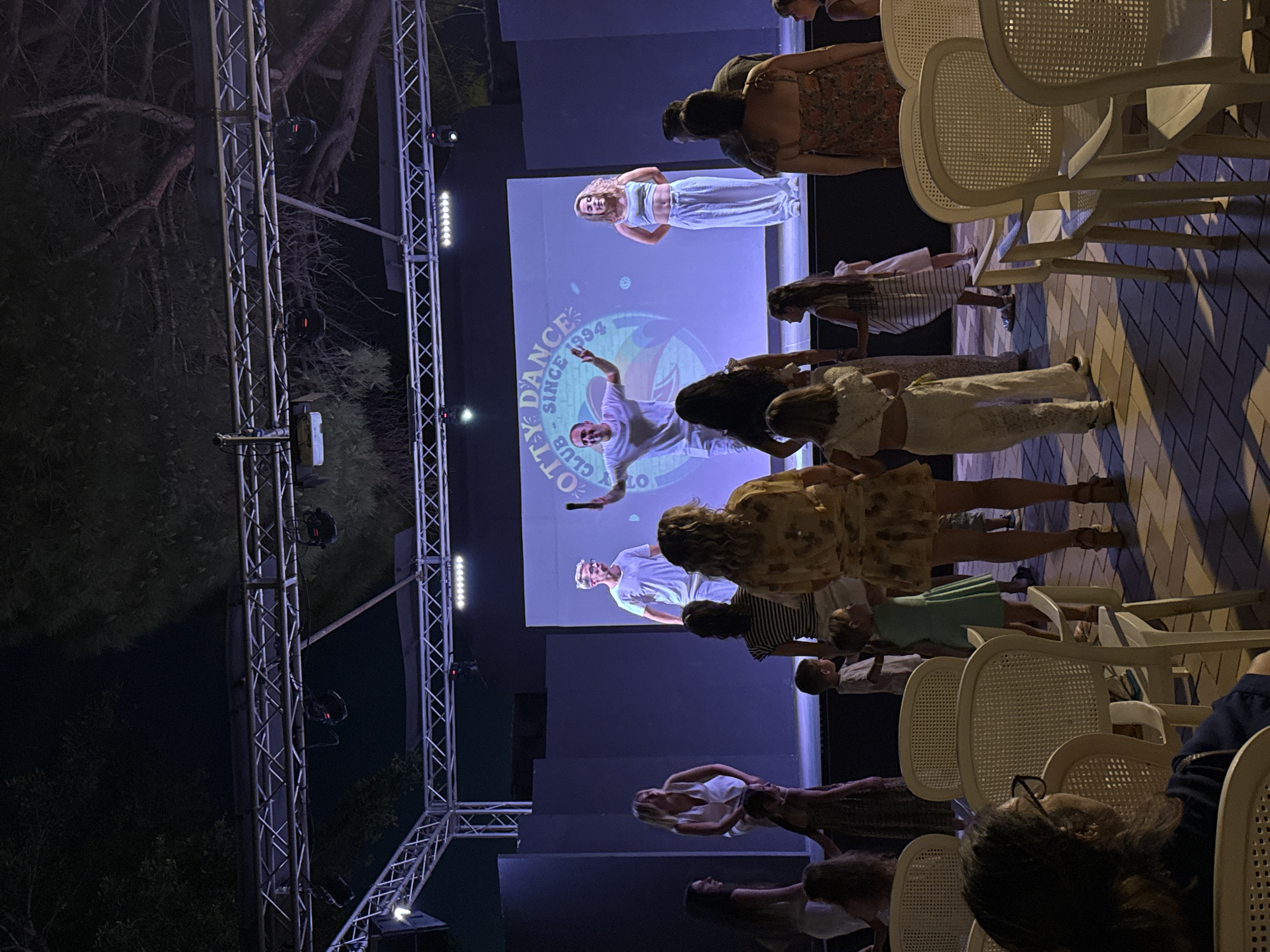 The image depicts an outdoor evening event at a location identified as being in Isola delle Femmine, Italy. A stage is set up with a large screen displaying a logo that reads "SPOTTY DANCE CLUB SINCE 1994". On the stage, two individuals are performing, a man in a white t-shirt and white shorts, and a woman in a white crop top and white pants. The man appears to be a performer or host, holding a microphone and gesturing. The woman stands next to him, facing the audience.
In front of the stage, a diverse group of people are gathered. Several adults and children are standing and watching the performance. Some individuals are seated in white chairs arranged in rows, facing the stage. These chairs have a cane-like woven back and seat. The ground in front of the chairs is paved with patterned tiles, casting shadows from the chairs and the people.
The background of the scene shows a dark, tree-filled area above the stage lighting rig, suggesting the event is taking place in a natural or semi-natural setting, possibly a park or resort. The stage is illuminated by various lights, creating a theatrical atmosphere. The overall impression is of a community or resort entertainment event.