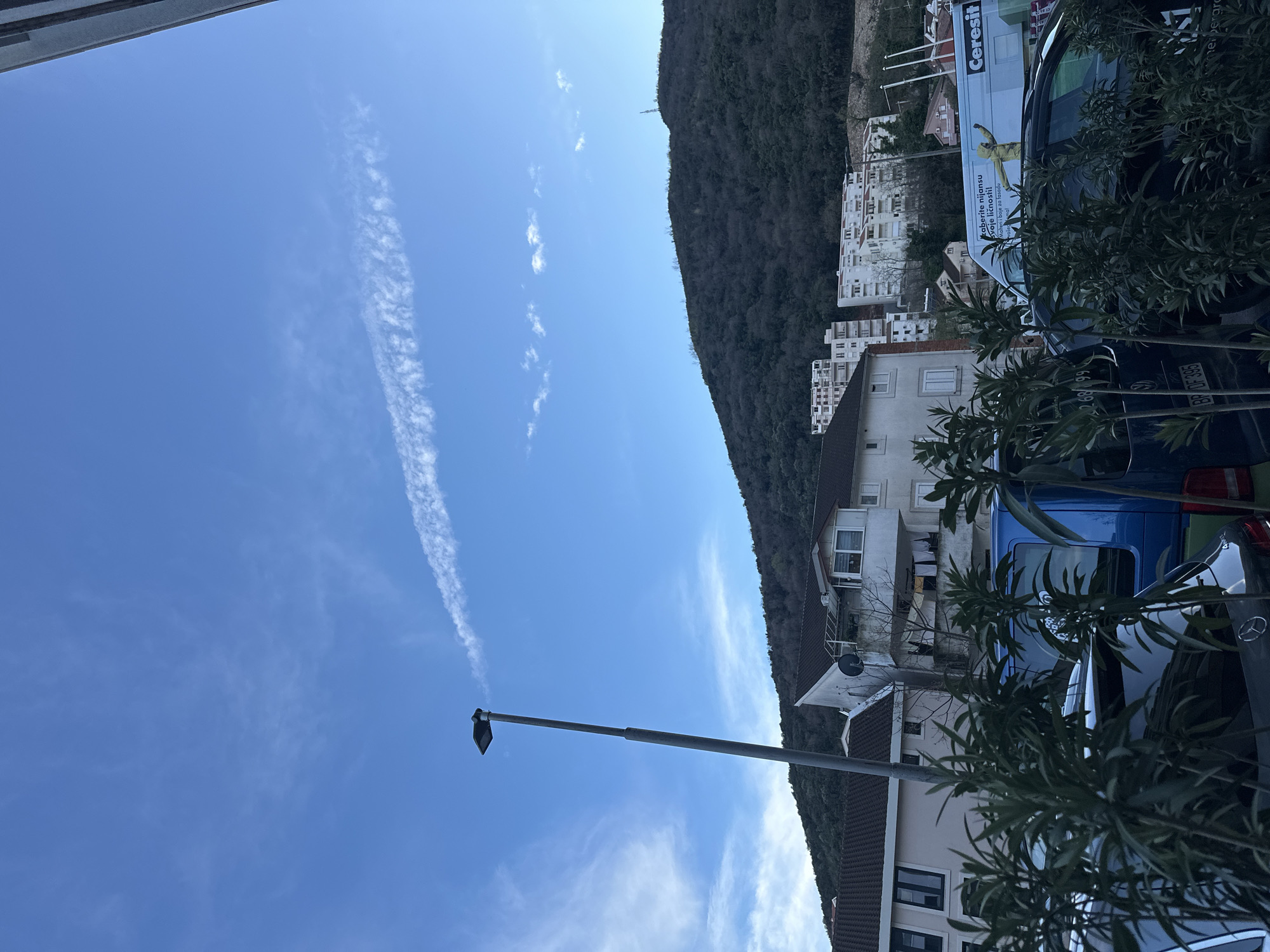 The image displays an outdoor scene in a residential area on a hillside in Budva, Montenegro. The sky is predominantly clear light blue, featuring a prominent, fragmented white contrail stretching horizontally across the upper middle, and sparse cirrus clouds in the upper left quadrant. A tall, gray streetlamp with a dark luminaire is positioned slightly left of center, extending from the lower mid-frame upwards.
In the foreground, dense green foliage partially obscures several parked vehicles. From left to right, a dark sedan with a visible Mercedes emblem on the trunk, a blue Volkswagen van with the Montenegrin license plate "BP OF 395" on its rear, and a white commercial van are visible. The white van is branded "Ceresit" and displays text in Montenegrin, including "Izaberite nijansu svoje ličnosti!" and "Mali i boje za fasade," along with a graphic of a person in a yellow protective suit. A white truck with "Jugoline" and a partial phone number ("067 268 991") is partially visible behind the Ceresit van.
Behind the vehicles and foliage, several residential buildings with light-colored facades and dark tiled roofs ascend the slope. One multi-story building in the mid-ground features multiple balconies, a satellite dish, and clothes hanging on a line. Further up the hillside, additional multi-story apartment blocks, predominantly white, are integrated into the landscape. The background is dominated by a large, dark green, heavily forested mountain. A communication tower or similar structure is discernible near the mountain's summit on the right side. No individuals are depicted in the scene.