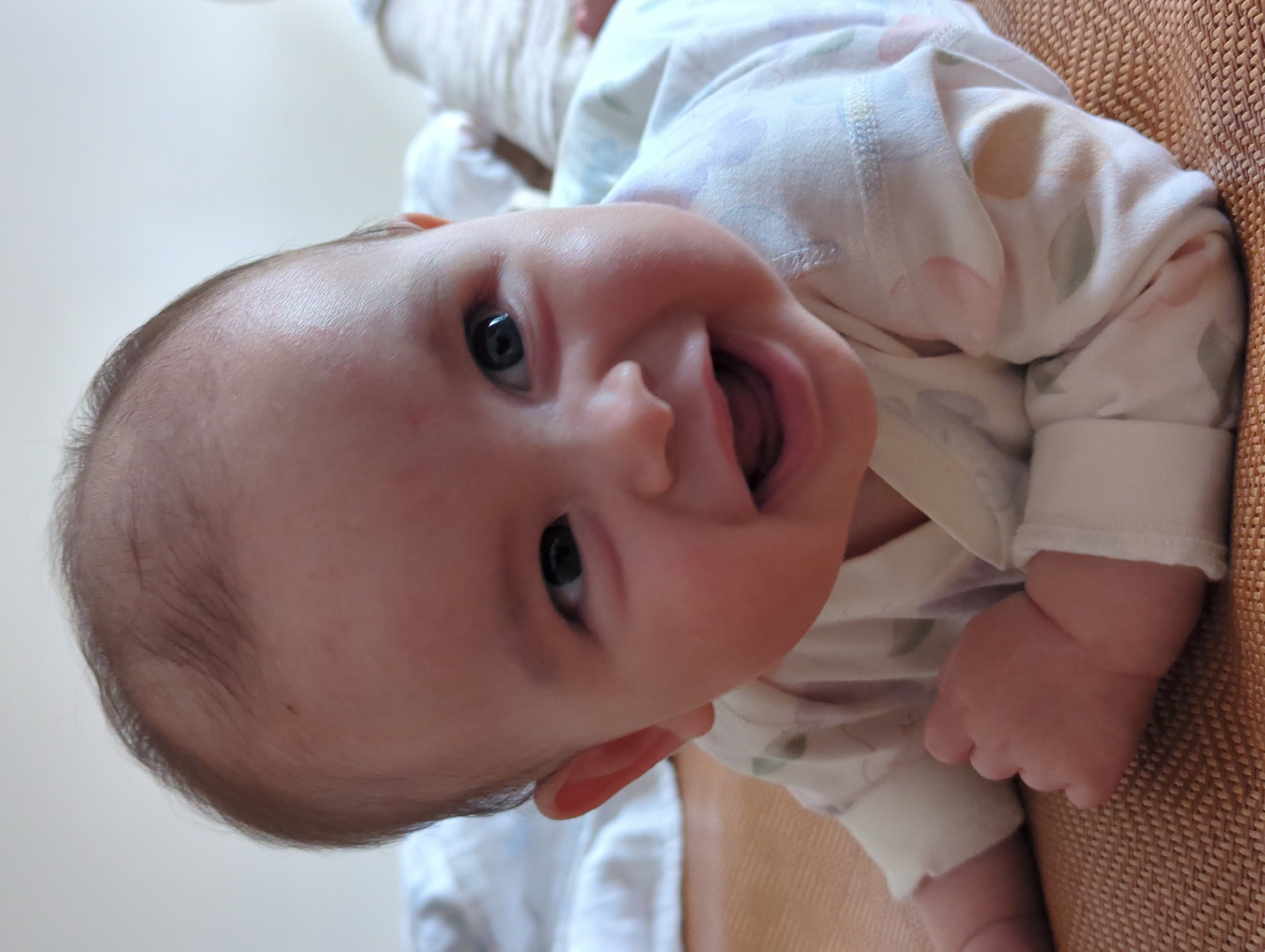 A close-up, tilted portrait shows a baby lying on their stomach, facing upwards with their mouth open and tongue visible. The baby has dark hair and light skin, with dark eyes looking directly at the camera. The baby is wearing a light-colored pajama top with a patterned design. The background is a blend of light beige on one side, possibly a wall, and a textured brown surface on the other, likely a couch or mat. No information in the image indicates a location in Shunde District, China.