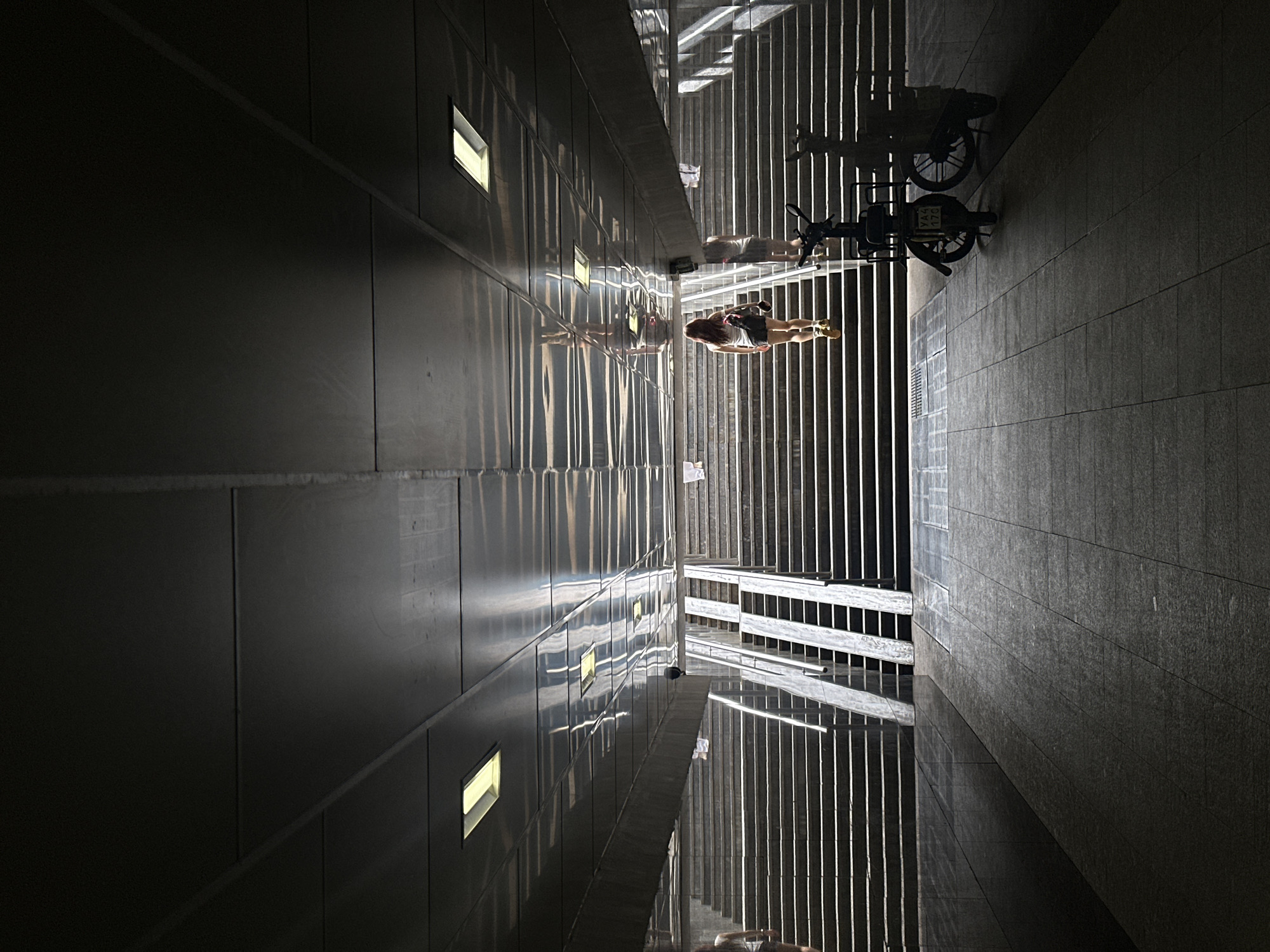 A slightly-rotated image depicts an outdoor scene with an urban aesthetic, likely a narrow alley or passage between two buildings. The scene is dominated by light and shadow.
On the left, a sheer wall with several square windows is visible. It reflects a set of vertical bars or railings in its surface.
In the center, a person in shorts and a t-shirt is shown. A bicycle with a basket is near the person.
On the right, a paved surface with noticeable tiles. The vertical bars are reflected in the pavement.