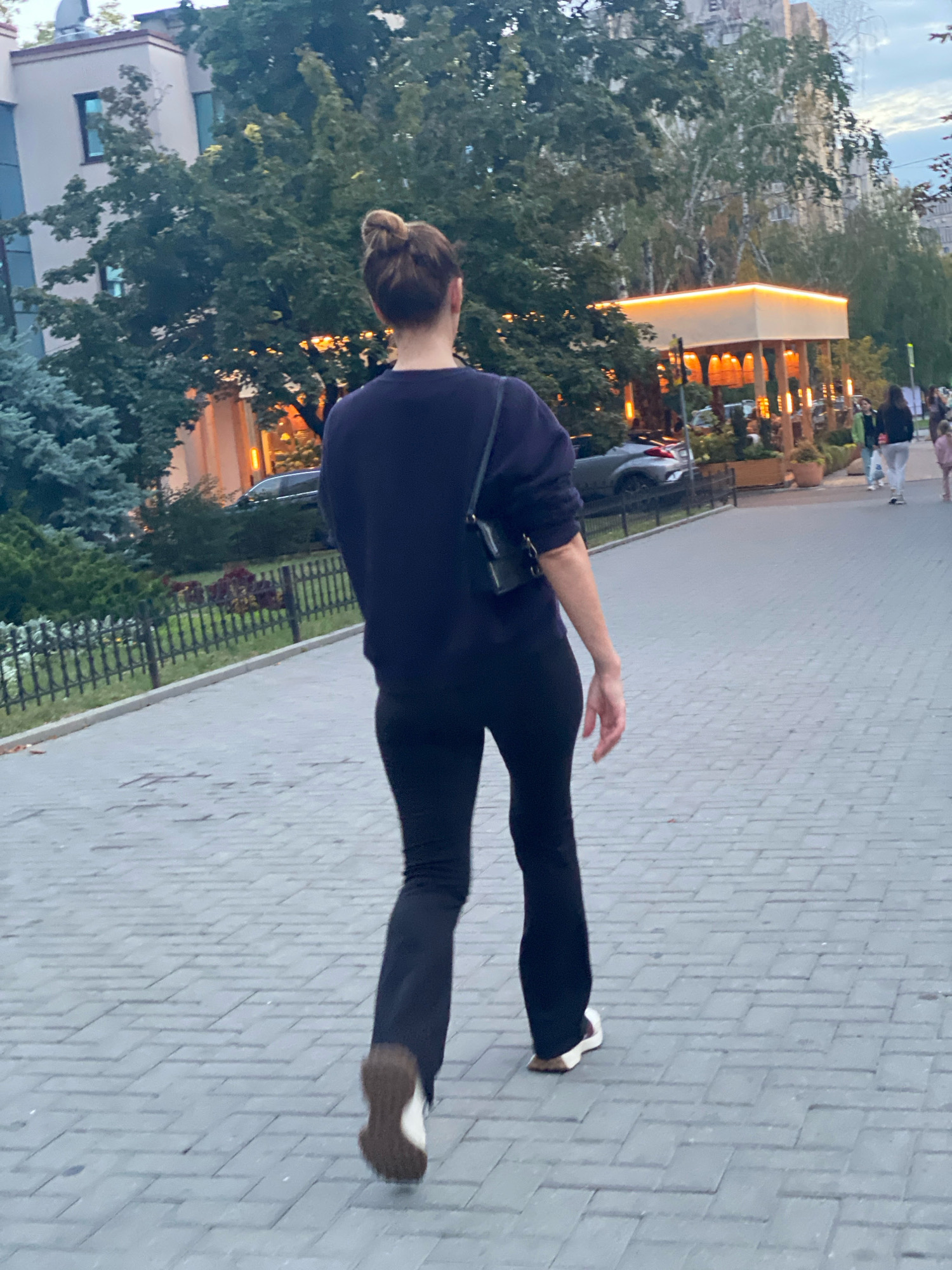 The image shows a woman walking away from the camera on a paved walkway. She has her hair styled in a bun and is wearing a dark long-sleeved shirt and dark flared pants. She is carrying a small dark crossbody bag. Her footwear appears to be athletic shoes with white soles and brown accents. The walkway is bordered by a black metal fence and green grass with some plants. In the background, there are trees, buildings, and a brightly lit outdoor seating area of a restaurant or cafe. Several other people are visible in the distance, walking on the same path or near the cafe. The environment suggests an urban setting, possibly a park or pedestrian area in Chișinău, Moldova, given the context provided.
