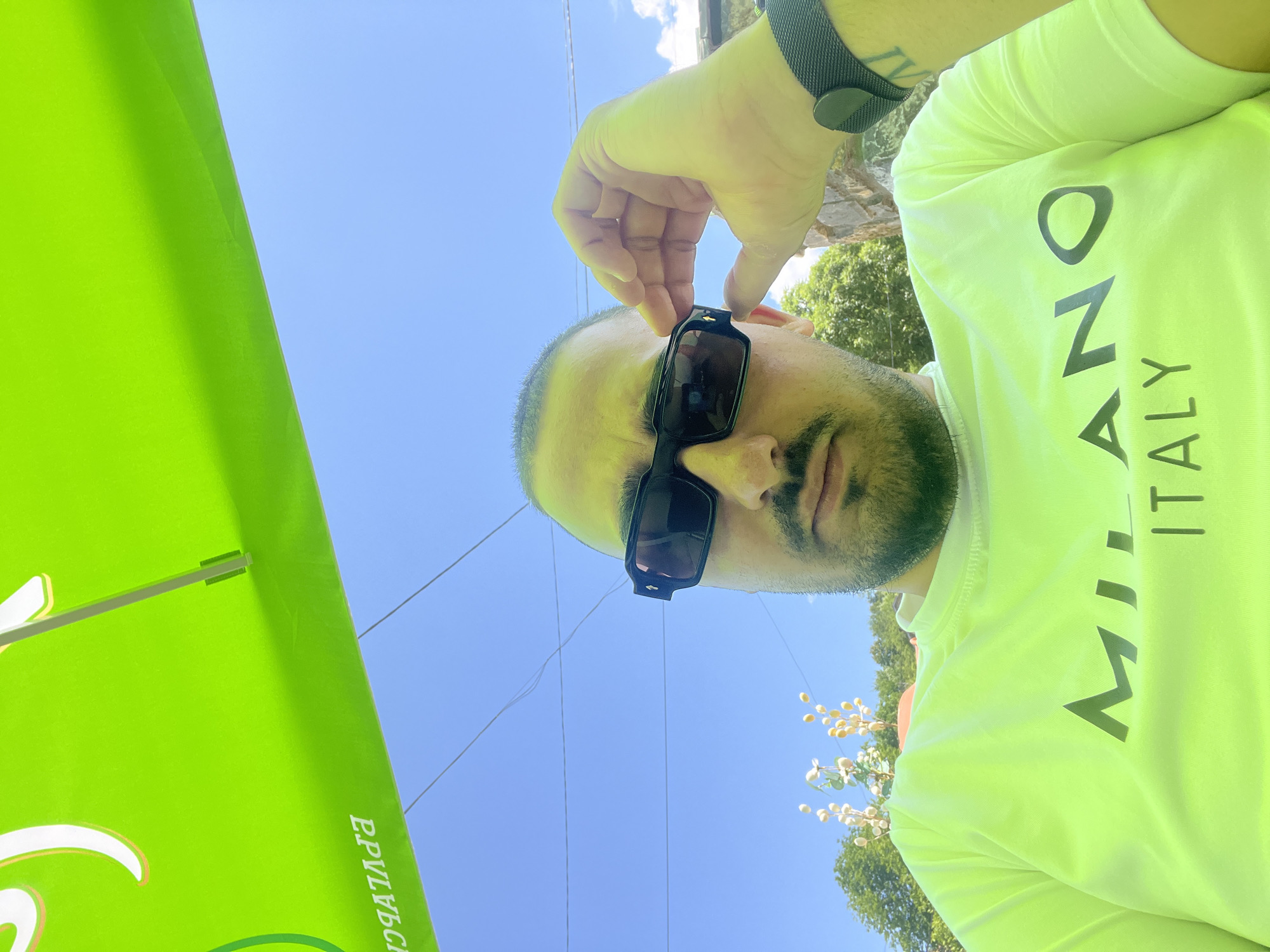 A close-up, low-angle portrait shot captures an adult male outdoors, identified as being in Касапите, Bulgaria. The man looks directly into the camera. He has short, dark hair, a beard, and a mustache. His skin exhibits a yellowish hue, consistent with illumination reflected from the large, bright green umbrella positioned directly above him, which occupies the top third of the frame. The umbrella's underside displays reversed white and orange/yellow Cyrillic text, reading "ЕРУЛЧВСН" (implying "НСВЛЧУРЕ").
The man wears dark, thick-framed rectangular sunglasses with a small, gold-colored detail on the outer edge of the right temple. His right hand (viewer's left) is raised, with fingers gently touching the right temple of his sunglasses, as if adjusting them. On his left wrist (viewer's right), he wears a dark, mesh-patterned wristband or watch, and a faint tattoo is visible on his inner forearm below the wristband. He is dressed in a neon green/yellow crew-neck t-shirt featuring the words "MILANO ITALY" printed in dark grey/black; "MILANO" is in a larger font above "ITALY."
The background reveals a clear, light blue sky traversed by several thin, dark horizontal lines, likely utility wires. Below the horizon line formed by the umbrella, green foliage from trees and bushes is visible. In the mid-ground, light-colored, decorative botanical elements resembling dried flowers or seed pods on slender stems are present. A light-colored stone or concrete structure is partially visible on the far right edge of the frame. The overall scene suggests a sunny day.