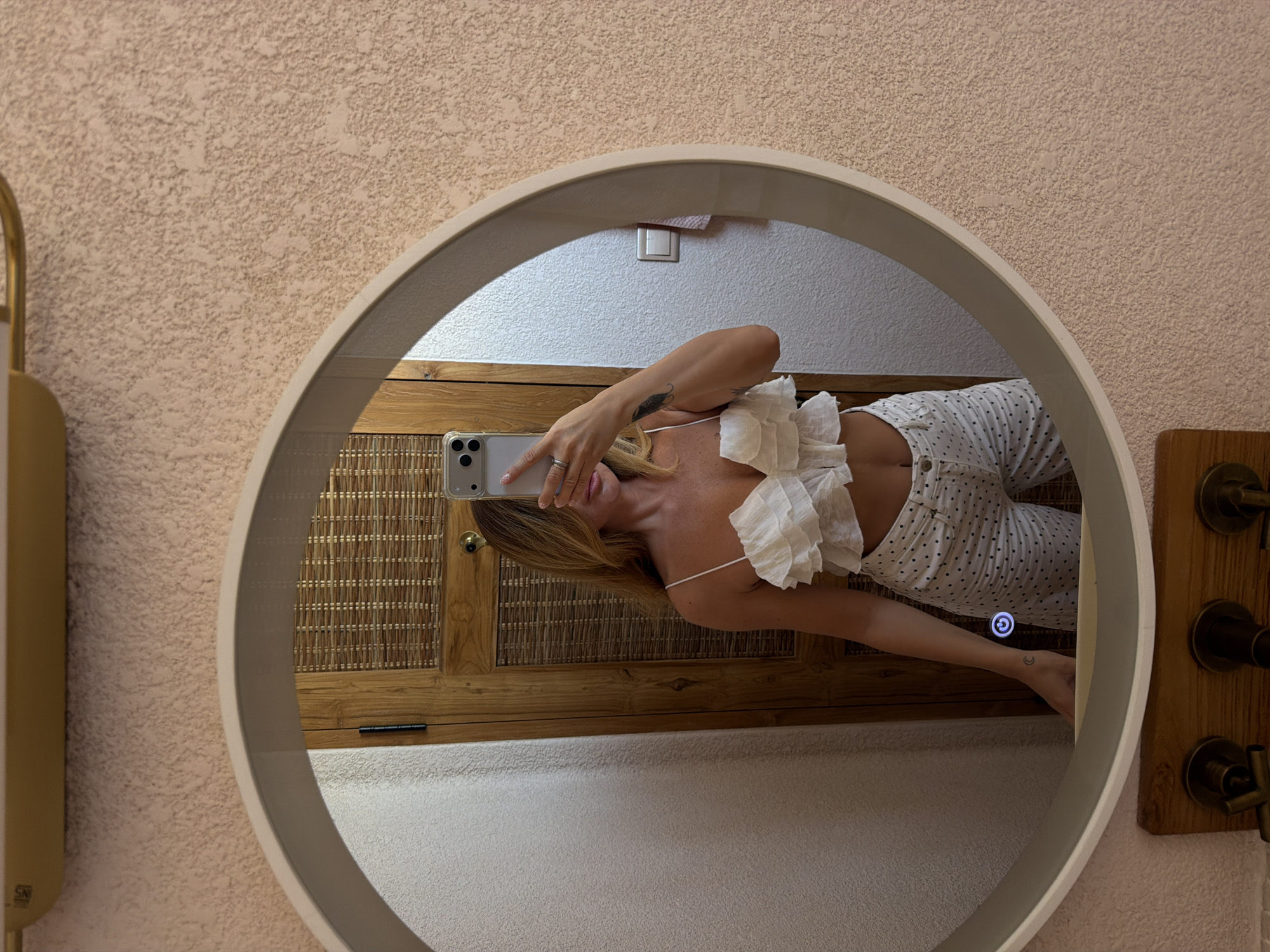 A woman is taking a selfie in a round mirror. She is wearing a white ruffled crop top and white pants with black polka dots. The mirror is hanging on a textured pink wall. Behind her in the reflection, there is a wooden cabinet and a light switch. Below the mirror, there are some brass faucets. The scene appears to be in a bathroom.
