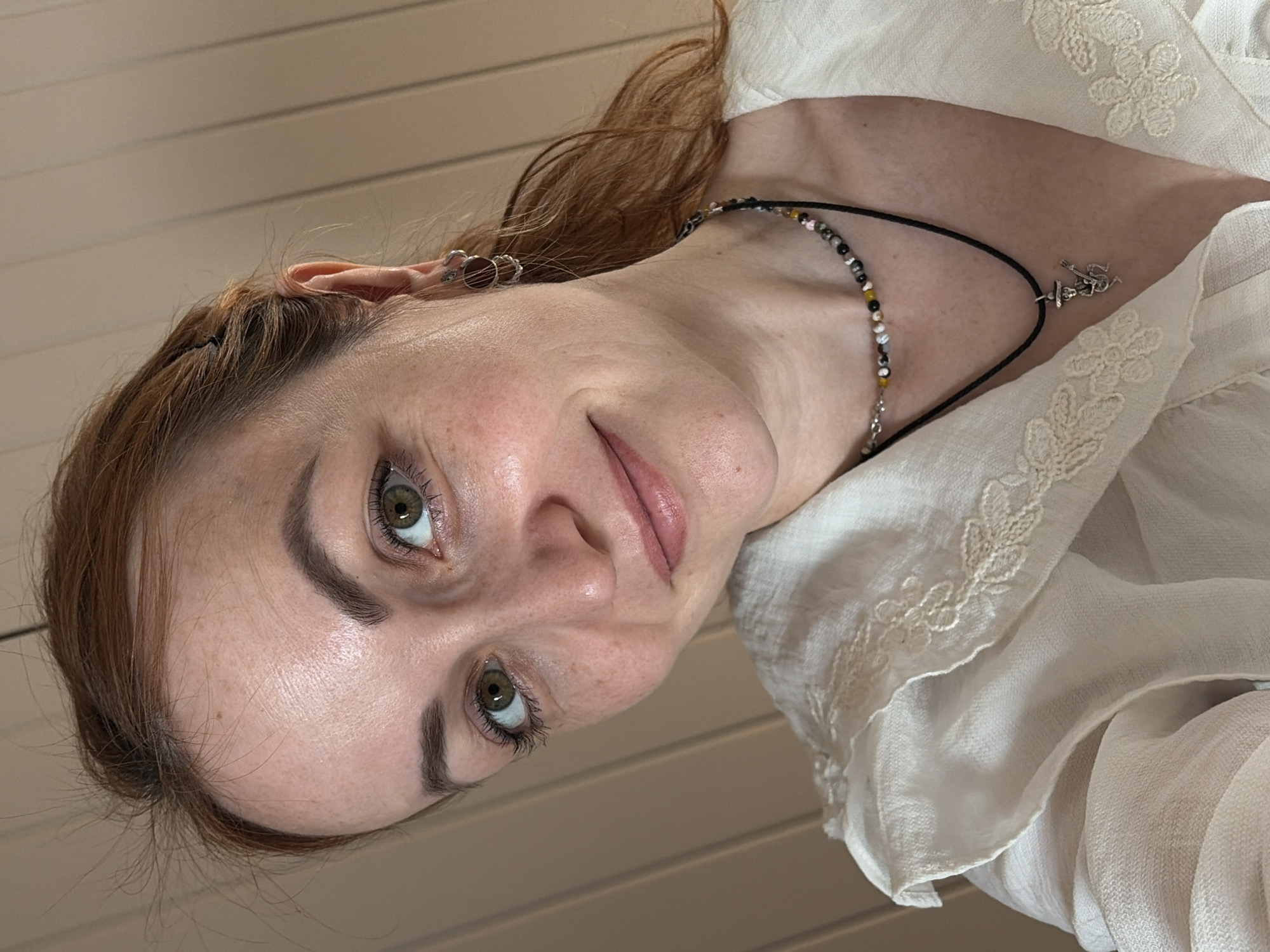 A selfie of a Caucasian woman with auburn hair and fair skin is presented. Her head is tilted to the right. She has freckles and green eyes. She is wearing a white blouse with lace trim and a black beaded necklace with a silver pendant. The background appears to be a light-colored wall or ceiling. The city of Chaikovsky, Russia, cannot be confirmed as the location based solely on the image provided.