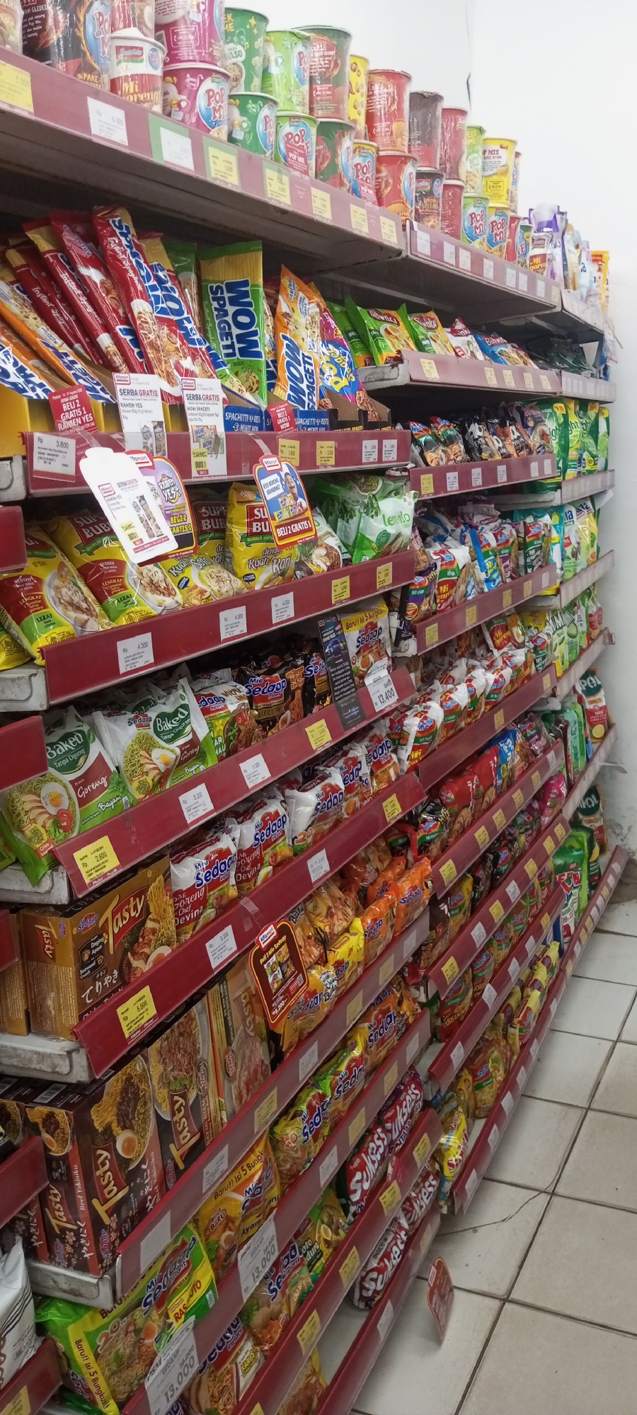 The image shows a supermarket aisle in Setu, Indonesia, filled with shelves of packaged food items. The products are predominantly instant noodles, snacks, and drinks, displayed in colorful packaging. The shelves are made of red metal, and price tags are visible on some items. The lighting appears to be from overhead fluorescent lights. The floor is tiled with a light-colored pattern. No people are visible in the image.