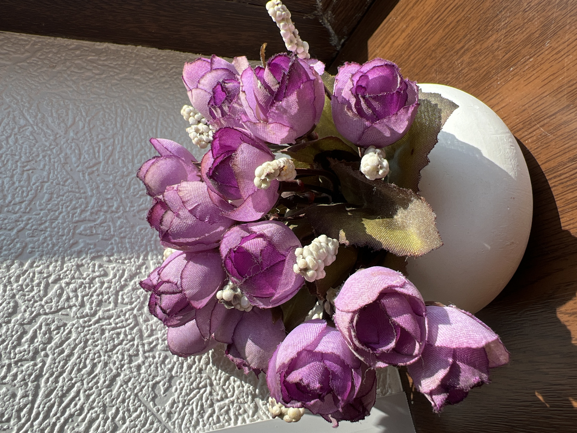 A bouquet of artificial purple roses is displayed in a white ceramic vase on a wooden surface, possibly a windowsill, in Marneuli, Georgia. The textured white wall behind the vase suggests an interior setting. Sunlight casts shadows on the wall and the wooden surface, indicating a bright day. The roses are fabric, with visible stitching and a slightly frayed appearance. Small white floral accents are interspersed among the larger blooms. The overall composition is still life, focusing on the arrangement of flowers and their interaction with light and shadow. There are no people or animals present.