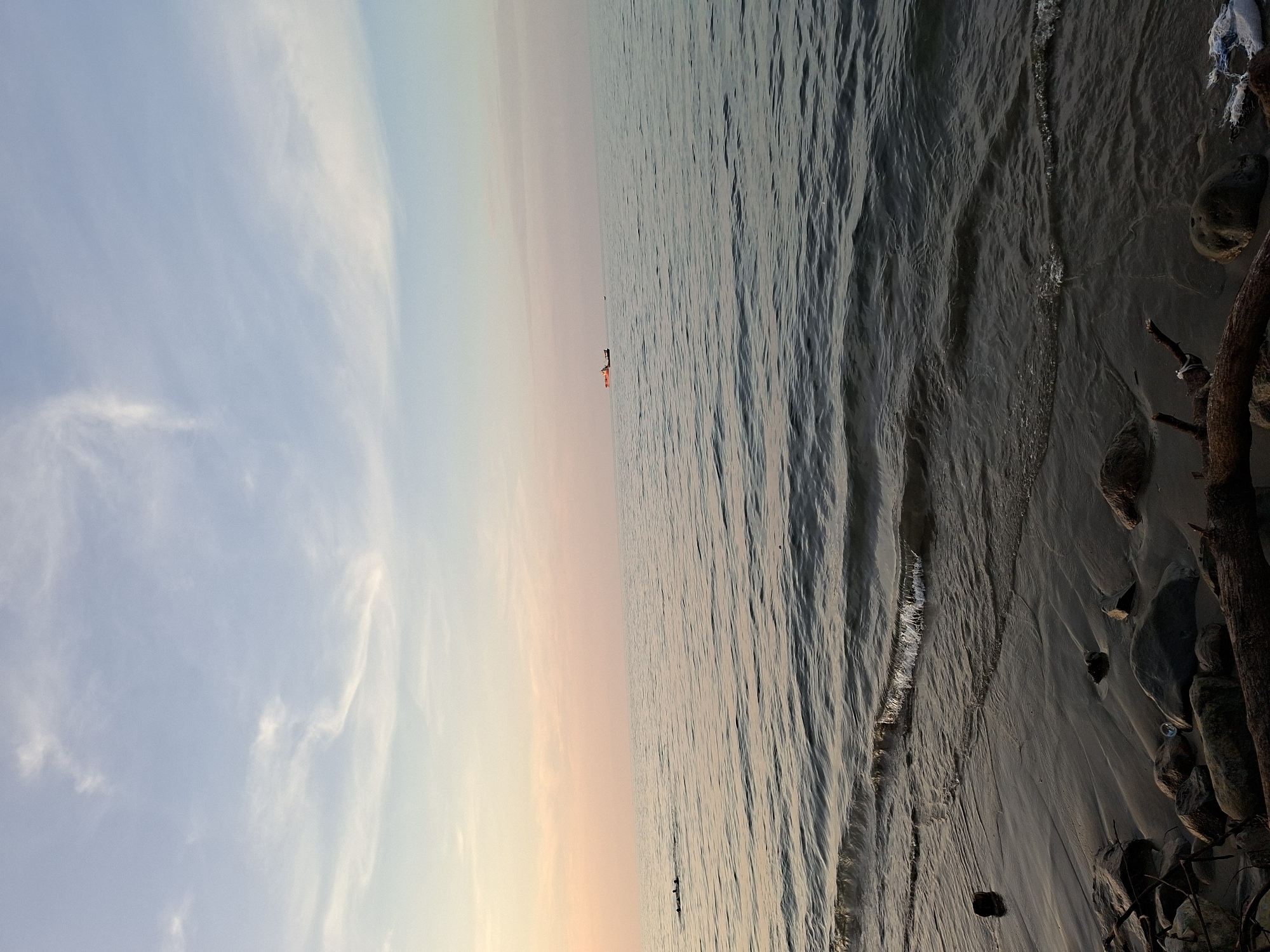 This serene image, captured in Lhokseumawe, Indonesia, depicts a coastal scene during either sunrise or sunset. The horizon line subtly divides the frame, showcasing a expansive sky above calm sea waters and a shadowed shoreline.
The sky presents a beautiful gradient of colors, transitioning from a light, clear blue at the top to soft peach and pale orange tones as it approaches the horizon. Delicate, wispy clouds are scattered across the bluer sections of the sky, adding depth and texture to the vast expanse.
Below the horizon, the tranquil sea stretches out, its surface marked by gentle ripples and small waves that reflect the muted hues of the sky. In the distant mid-ground, two small figures are visible on the water. One appears to be an orange kayak or small boat with a person inside, accompanied by another smaller, darker shape nearby, suggesting a peaceful recreational activity such as paddling or fishing.
The immediate foreground on the right reveals a dark, somewhat rugged shoreline composed of sand and rocks. Elements of driftwood or tree roots are present, alongside what appears to be discarded white plastic or fabric debris. The water meets the land gently, indicating calm conditions. The overall atmosphere of the image is one of profound peace and tranquility, highlighting the natural beauty of the environment during a quiet time of day. No text is visible in the media file.