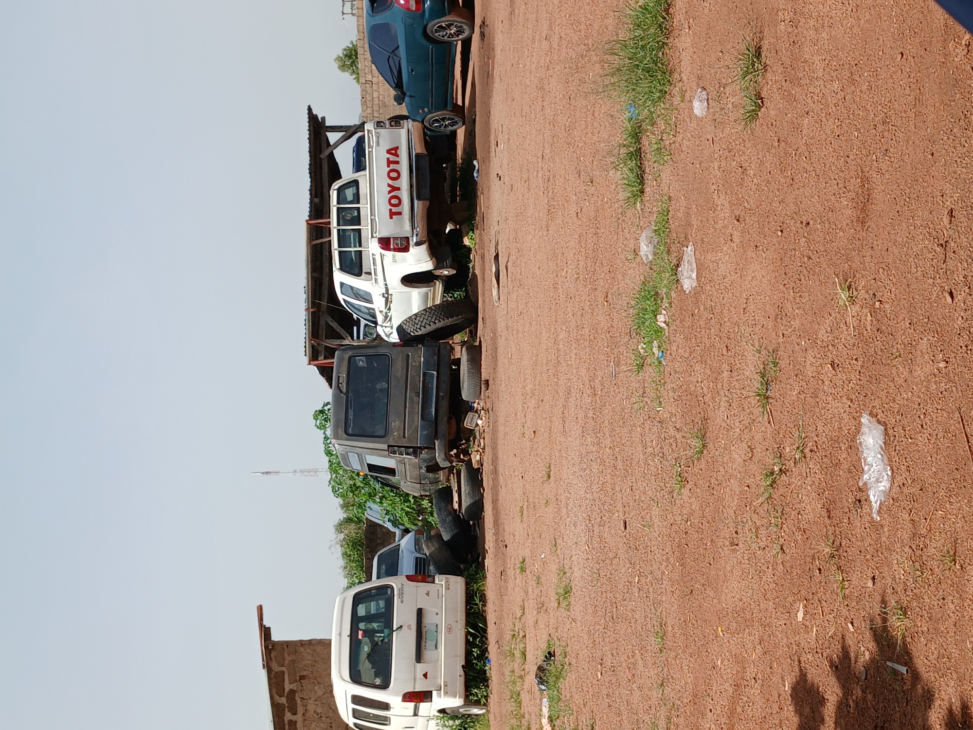 The image depicts an outdoor scene in Gura Top, Nigeria, likely a vehicle repair or storage lot. Several vehicles are present, including a white van on the left, a dark-colored van in the center, a white Toyota pickup truck in the middle, and a dark green sedan on the right. The Toyota truck has "TOYOTA" written in red on its tailgate. Tires are scattered around the vehicles, some piled up near the dark van and others lying on the ground. A simple wooden shelter with a corrugated roof is visible behind the Toyota truck. The ground is a reddish-brown dirt, with patches of dry grass and some scattered litter, including plastic bags. A low brick wall and some trees are visible in the background, along with a distant cell tower. The sky is clear and pale blue. No people are visible in the image.