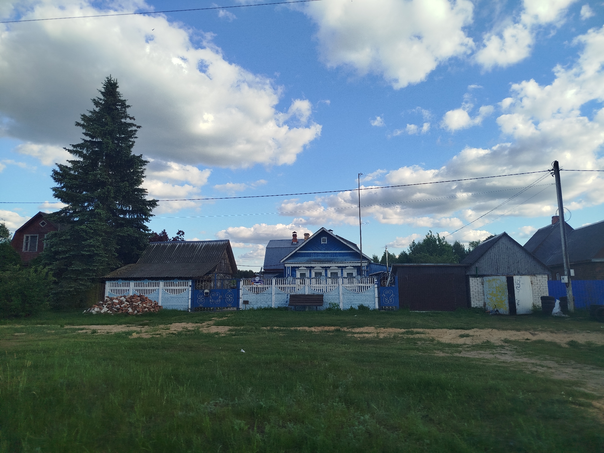The image shows a rural scene in Заречье, Russia, under a partially cloudy sky. A blue wooden house with ornate window frames and a white picket fence is centrally located. Adjacent to the blue house are smaller wooden structures, including a dark grey building with a gabled roof to the left and a light grey brick structure with a sliding door to the right. An evergreen tree stands tall to the left of the blue house. In front of the house and structures, there is a grassy field with dirt paths. Power lines are visible against the sky. A pile of bricks is positioned near the left side of the image.