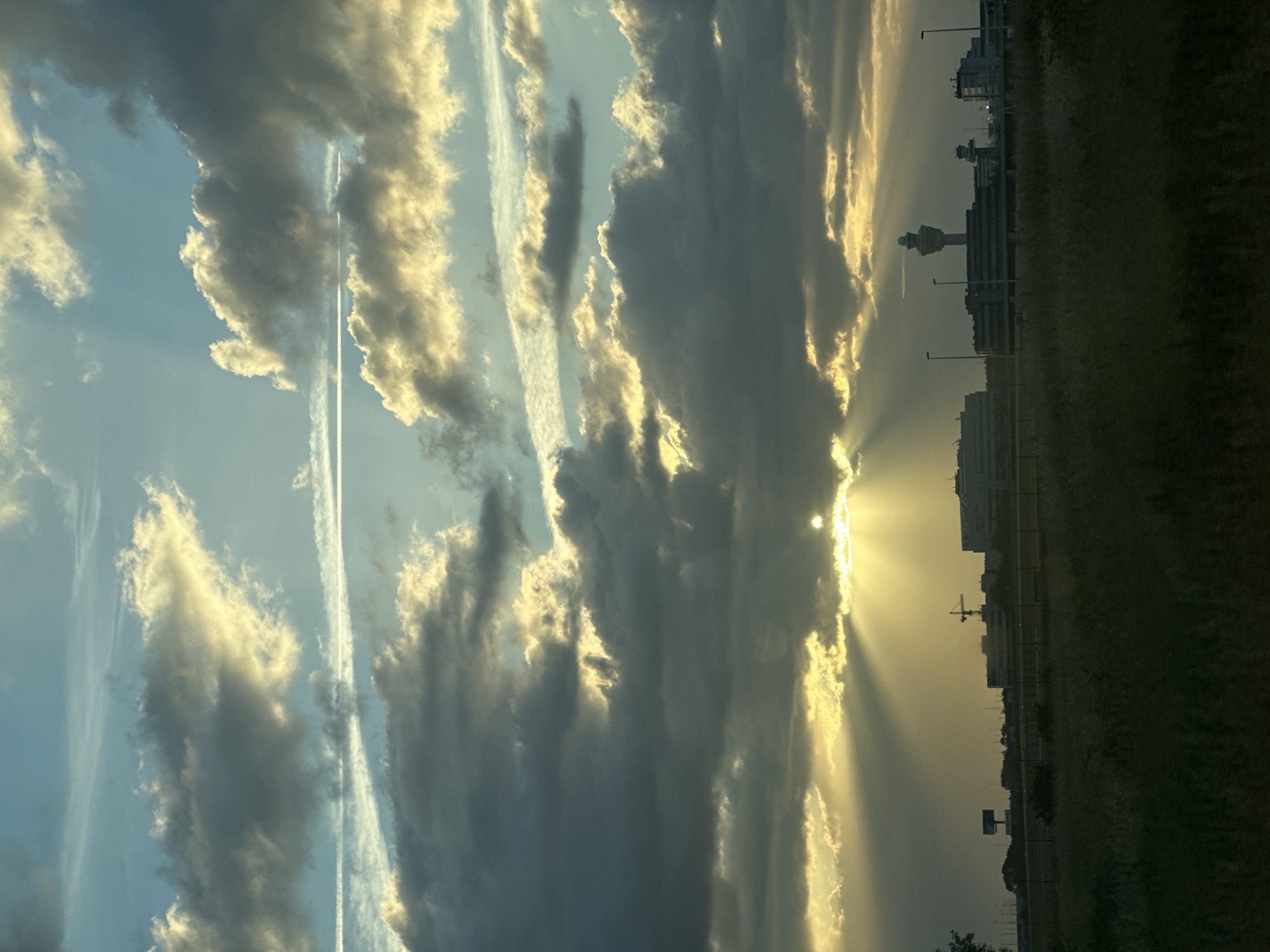 The image depicts a dramatic sunset over a landscape that includes an airport control tower and other airport buildings, consistent with the location of Schiphol Airport near Hoofddorp, Netherlands. The sky is filled with a mix of dark, ominous clouds and bright, sunlit clouds, with visible sunbeams piercing through the darker formations. The sun itself is partially obscured by clouds, casting a warm, golden light. In the foreground, there is a grassy field with some indistinct vegetation. The middle ground features a line of airport structures, including a prominent control tower, illuminated buildings, and what appears to be a construction crane. The horizon is marked by the silhouette of these buildings and some distant trees. The overall impression is one of a striking natural phenomenon occurring over a man-made infrastructure. There are no people visible in the image.