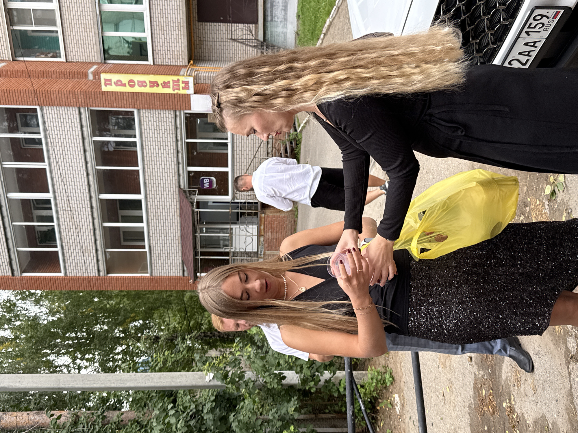 In an outdoor urban setting, two women and a man are visible. One woman, with long blonde hair and wearing a black dress with a sequined skirt, is holding a small clear cup with a pink drink. The other woman, also with long blonde hair and wearing a black top, appears to be taking the cup from her. The man, in the background, is wearing a white shirt and black shorts. The scene takes place near a brick building with multiple windows, and a yellow sign with Russian text hangs vertically on the facade. A white car with a license plate reading "2AA 159 RUS" is also present. The ground is paved with dirt and some fallen leaves, and a black metal railing is in the foreground. Trees and greenery are visible in the background, suggesting a slightly overcast day.