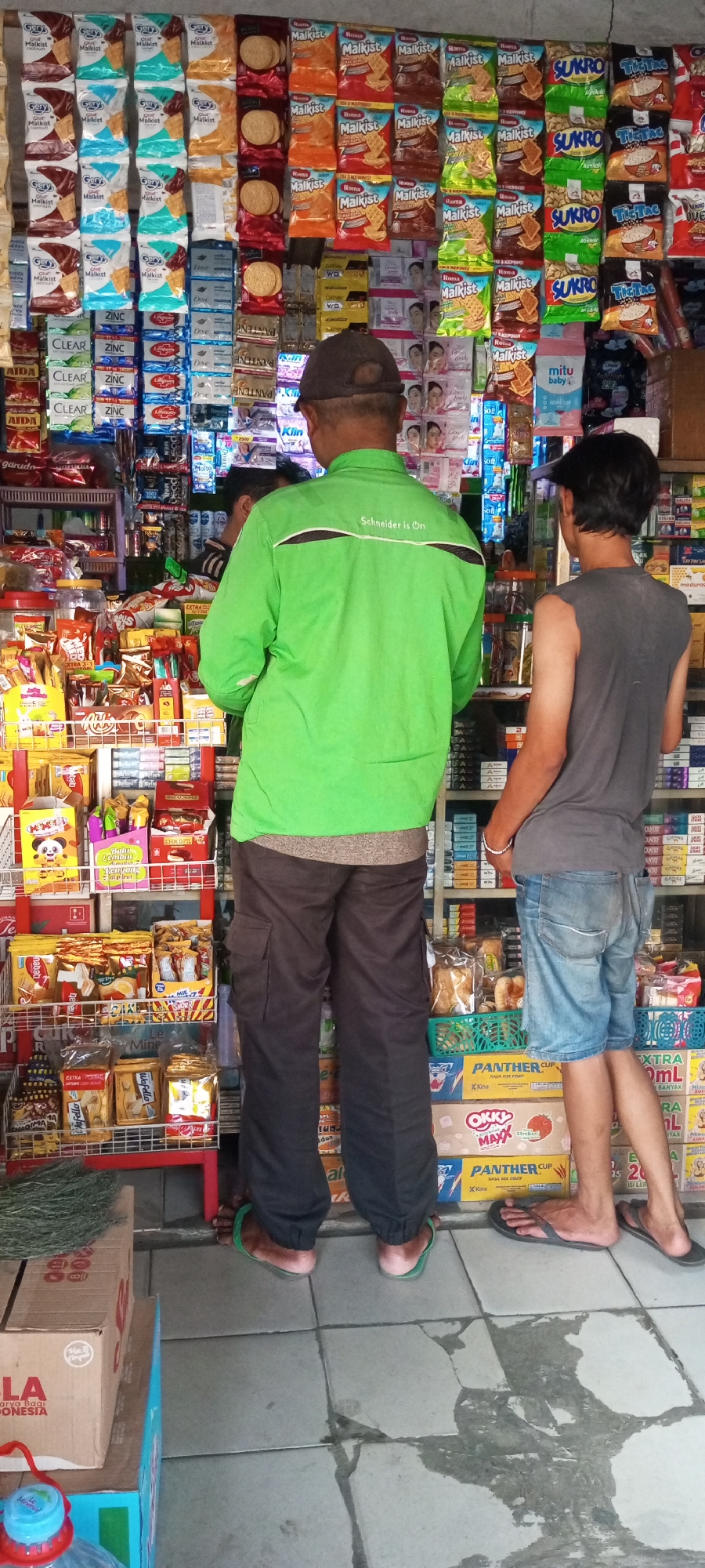 In Setu, Indonesia, two individuals are seen standing in front of a store stocked with various packaged goods. The person on the left, facing away from the camera, wears a bright green long-sleeved jacket with "Schneider is On" printed in white on the back, and dark cargo pants. The person on the right, also facing away, wears a dark gray sleeveless t-shirt and denim shorts. The store's interior is visible, displaying an array of snacks, beverages, and personal care items on shelves. The floor is tiled and appears wet in places. A cardboard box with Indonesian writing is partially visible on the left, and a blue plastic container with a red lid is on the floor in the foreground. The overall impression is a typical street-side shop scene.