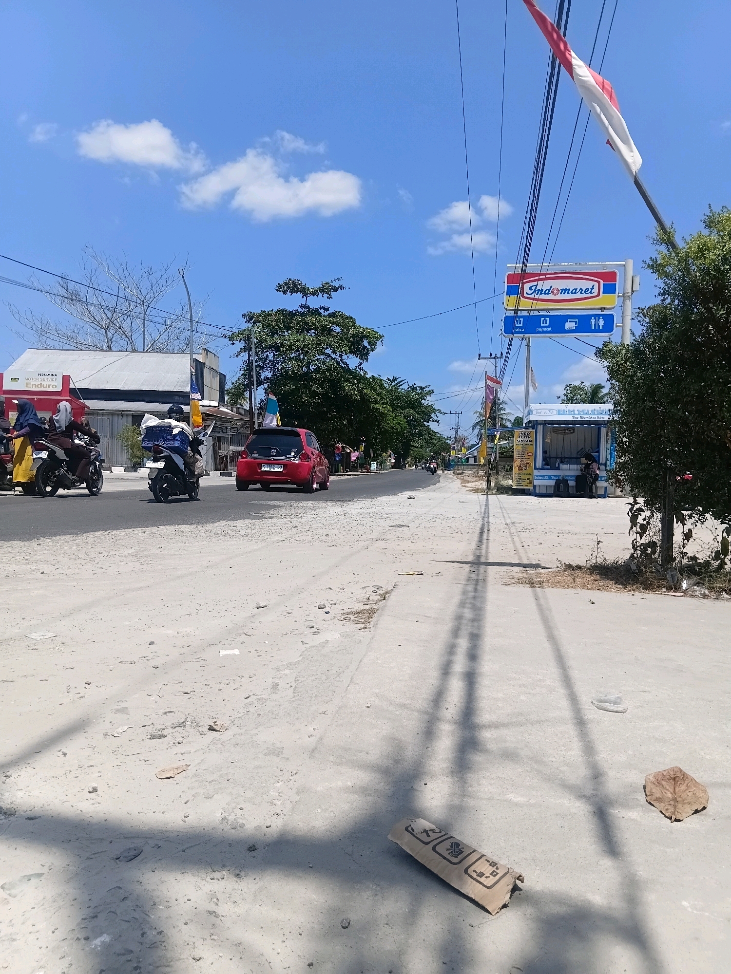 The image depicts a street scene in Sungai Kapitan, Indonesia, under a bright blue sky with scattered white clouds. The road in the foreground is dusty and unpaved, with some debris including a crumpled brown cardboard tube. To the right, a paved sidewalk runs alongside the road.
On the left side of the street, a red car is parked. Further down the road, several motorcycles are visible. One motorcycle, carrying a person wearing a red hijab and another person, is moving away from the camera. Another motorcycle, with a driver wearing a helmet and carrying a large white package on the back, is also moving away. In the distance, a building with a white roof and metal siding is visible, with the signage "PERTAMINA MOTOR SERVICE ENDURO" on its side.
On the right side of the street, a blue and white kiosk-like structure, likely a convenience store, stands with a sign that reads "INDOMARET". An Indonesian flag is flying from a pole above the Indomaret sign. Two individuals are seated in front of the kiosk. Overhead, power lines crisscross the sky, casting shadows on the ground. A large green tree stands to the far right of the image, partially obscuring the view of the sky.