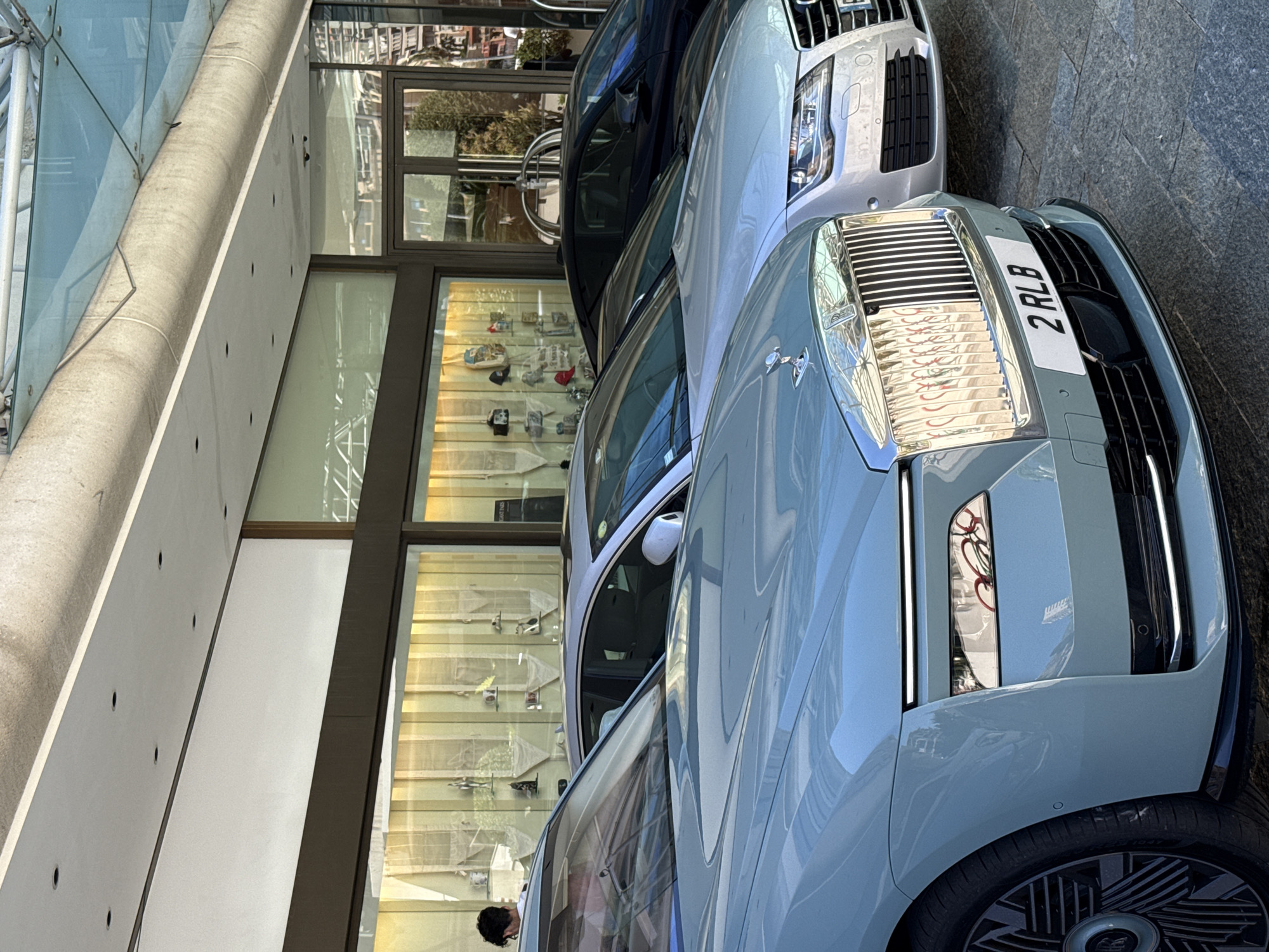 The image shows a row of luxury cars parked in front of a building with large glass windows, likely a high-end establishment in Monaco, Monaco. In the foreground, a light blue Rolls-Royce with a prominent chrome grille and the license plate "2 RLB" is visible. Behind it, a silver Audi sedan is parked, followed by a dark-colored car, possibly another luxury sedan or coupe. The building behind the cars features a well-lit display window filled with various items, suggesting it might be a boutique or a showroom. The ground appears to be paved with stone tiles. The overall scene conveys an atmosphere of wealth and sophistication, characteristic of the city of Monaco.