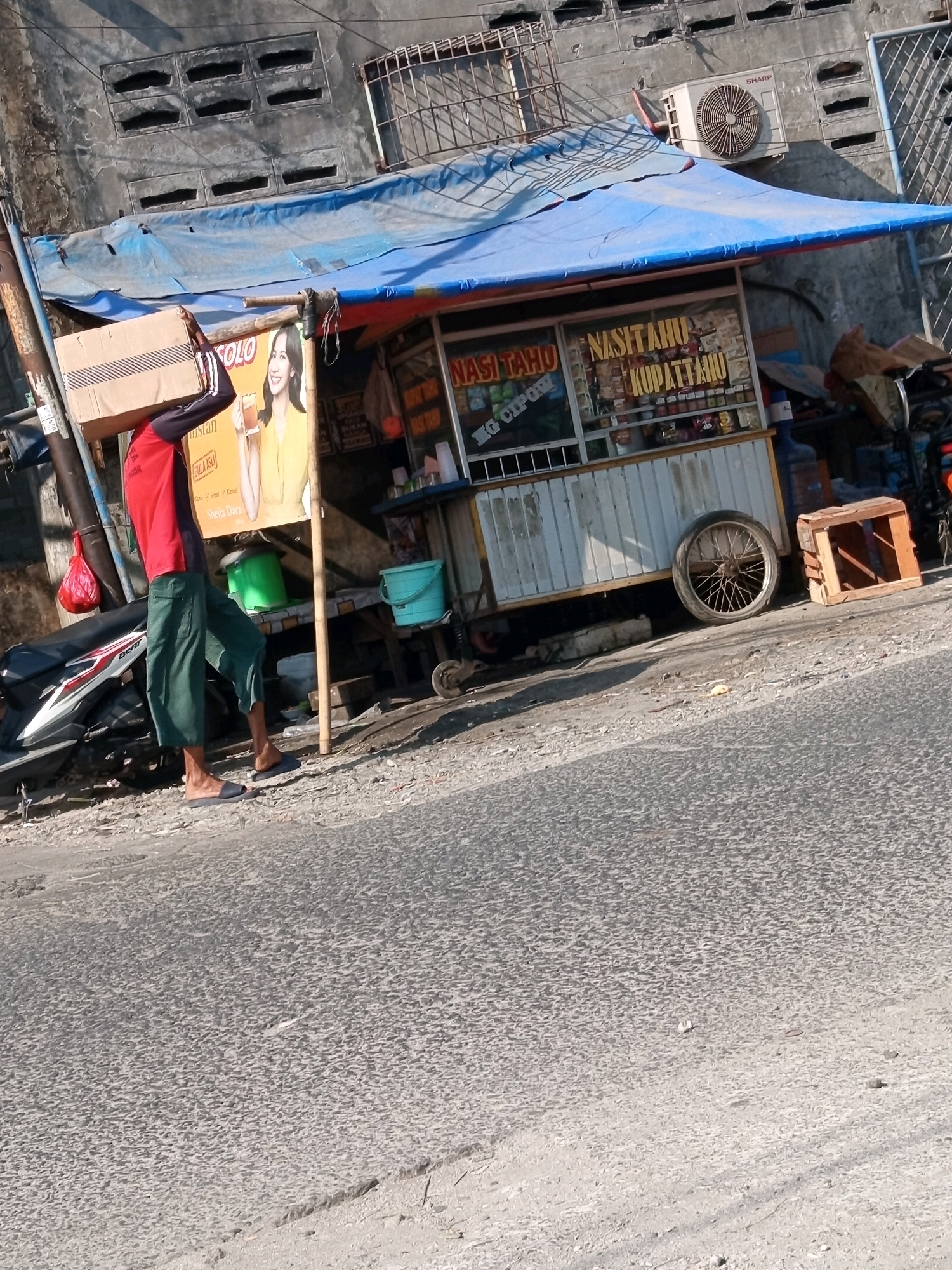 A man wearing a red shirt and green pants carries a cardboard box on his head, walking past a food stall in Karawang, Indonesia. The stall, covered by a blue tarp, displays various food items and beverages. A motorcycle is parked to the left of the stall. The scene is set on a roadside, with a paved street in the foreground and a building with a window air conditioning unit in the background.