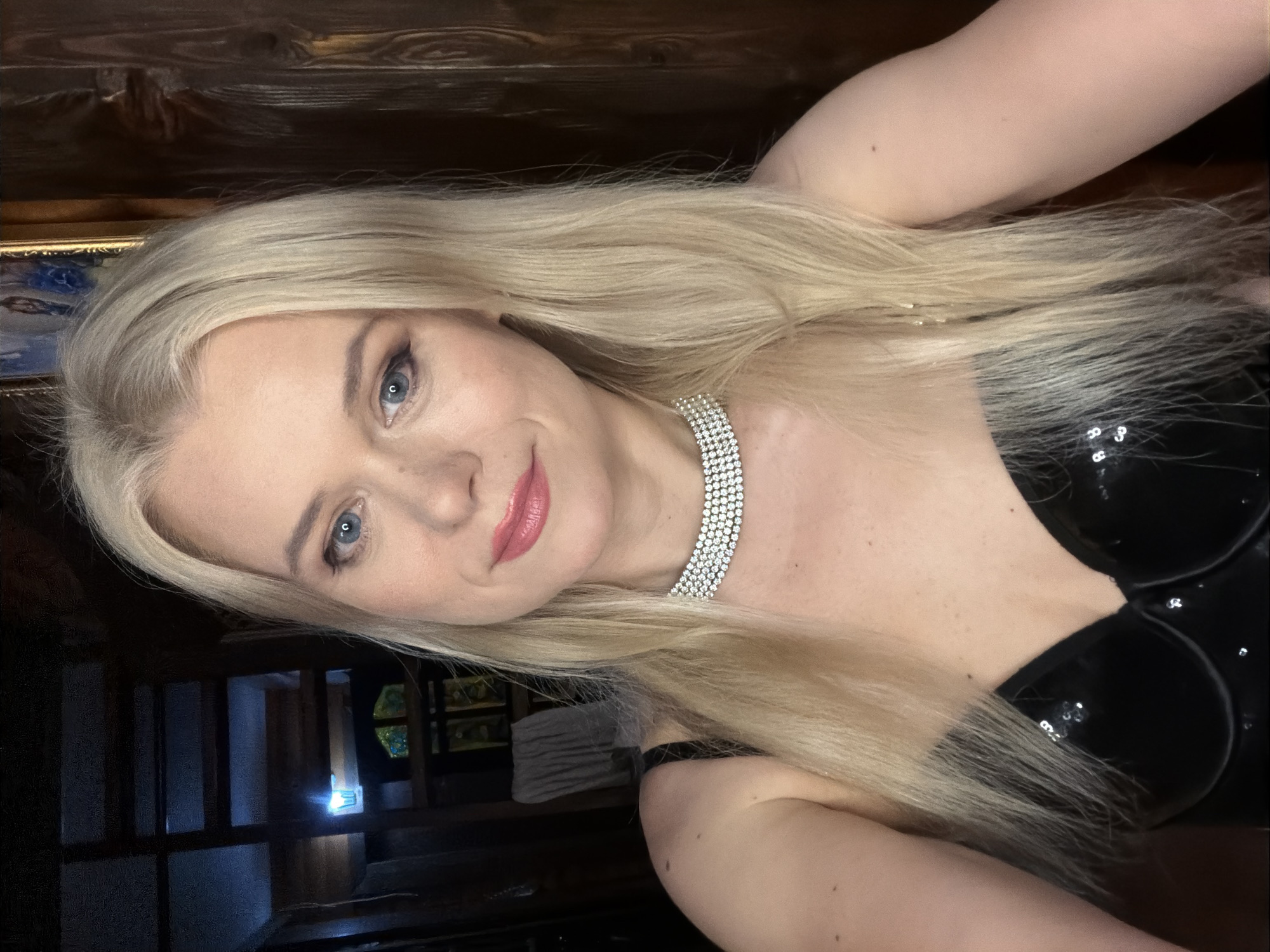 A blonde woman with blue eyes and pink lipstick poses for a selfie indoors. She is wearing a black sequined bustier and a diamond necklace. The background appears to be a rustic wooden interior with a staircase and stained glass. There is no indication of the city of Sochi, Russia, in the image.
