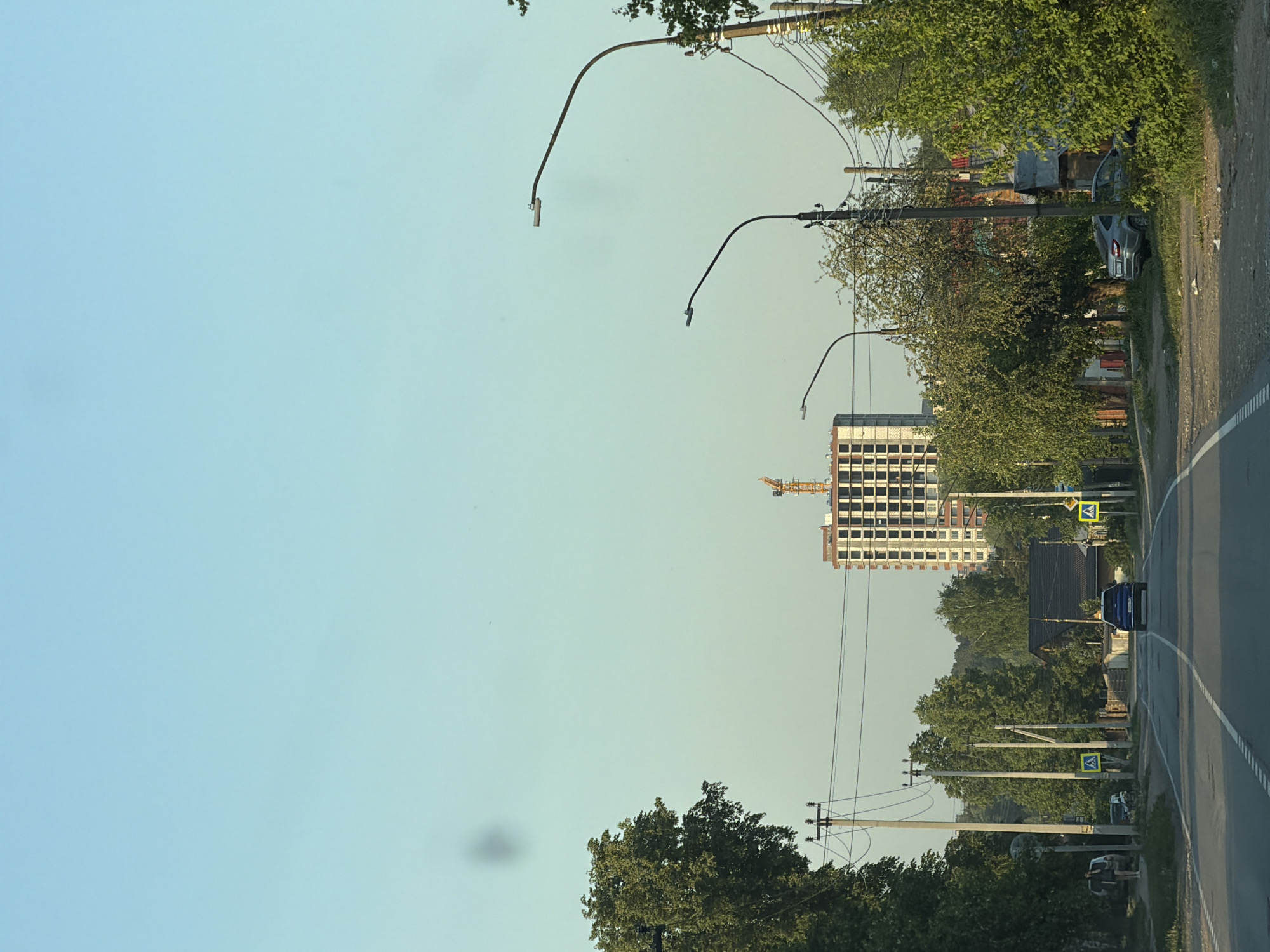 The image depicts a street in Chaikovsky, Russia, with a view of a building under construction. A road with white lane markings runs along the right side of the frame. A dark blue vehicle travels along the street. Overhead power lines connect utility poles on both sides of the road. Lampposts with curved arms are spaced along the street. A tall building under construction with a yellow crane is visible in the distance. Trees line the street. Pedestrian crossing signs are on the right side of the street. A light-colored car is parked on the side of the road on the right, partly obscured by vegetation. The sky is clear and light blue.