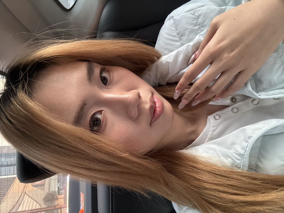 A young East Asian female is positioned from the chest up, seated in the rear passenger compartment of a vehicle, appearing to face the camera directly. She has long, light brown hair with darker roots, parted on the side. Her eyes are dark, and her facial expression is neutral. She is wearing a light-colored, possibly pale blue or green, quilted puffer jacket over a white, ribbed top fastened with metallic snap buttons. Her left hand is visible, resting on her chest or jacket, with fingers slightly spread. Her fingernails are long, manicured, and feature a light-colored base with small, dark speckled patterns. The background, visible through the vehicle's window, depicts an urban environment consisting of multi-story buildings with visible windows and several bare trees, indicating a daytime setting. This scene is situated in the city of Liangjiang, China.