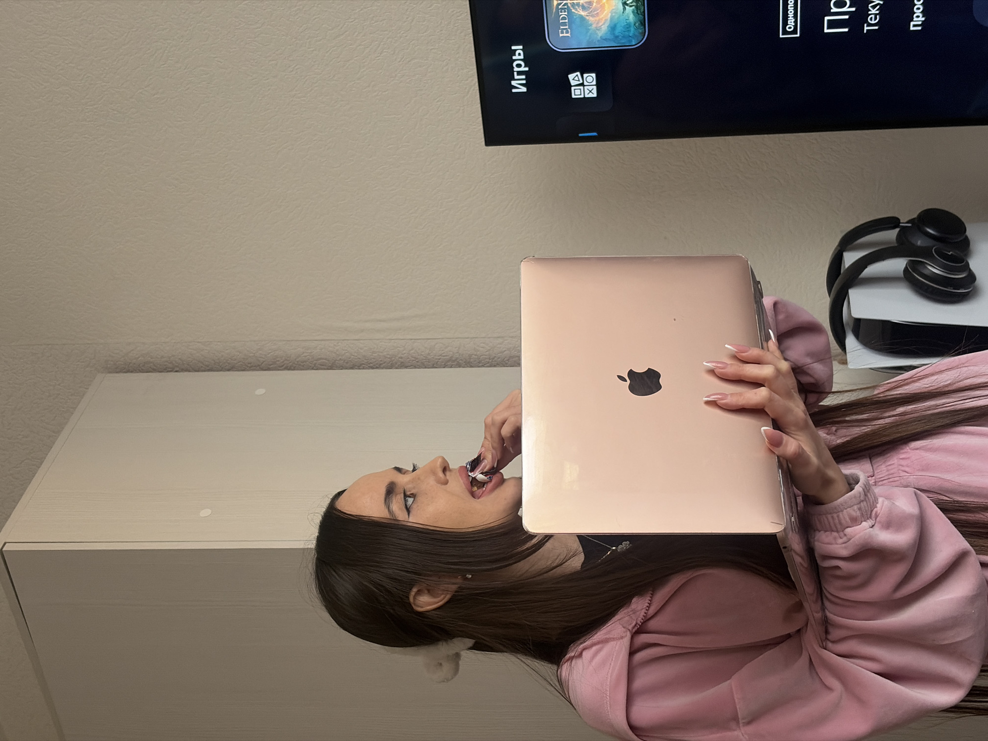 The scene is set indoors, within what is described as a room in Saint Petersburg, Russia, indicated by Russian text visible on a television screen. A young female individual with long, dark brown hair, tied back partially with a white scrunchie, is positioned in profile, facing right. She wears a pink velour tracksuit over a black top, accessorized with a four-leaf clover pendant necklace and small earrings. Her fingernails are manicured with French tips. She holds an open rose-gold Apple MacBook laptop in both hands, angled upwards. Her right hand is actively placing a small, white-wrapped object into her open mouth. To the right, a dark-screened television displays a menu, with the word "Игры" (Games) prominently at the top, PlayStation controller icons, and the cover art for the video game "Elden Ring." Additional Russian text, "Однопользовательская" (Single player), "Про" (About), "Текущая" (Current), and "Просмотр" (View), is visible below. Below the television, a white PlayStation 5 gaming console stands vertically, with a pair of black over-ear headphones resting on its side. The room features plain, light-colored, textured wallpaper, and a light-colored wardrobe or cabinet is partially visible on the far left.