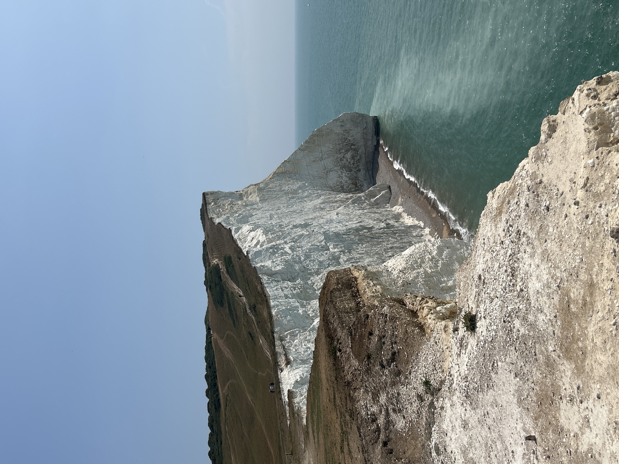 The image depicts a coastal landscape with dramatic white chalk cliffs meeting the turquoise sea. The cliffs are steep and sheer, with visible strata of chalk rock. A narrow strip of pebbly beach is visible at the base of the cliffs. The water is clear and appears to have varying shades of green and blue. On top of the cliffs, there is a grassy, undulating terrain with several dirt paths winding across it. A few small figures of people can be seen walking along these paths, and in the distance, a faint white sailboat is visible on the horizon. The sky is clear and a pale blue, with a few distant birds visible as tiny specks. The foreground of the image shows a rocky, eroded cliff edge with sparse vegetation. This scene is characteristic of the Seven Sisters cliffs near Seaford in East Sussex, United Kingdom.