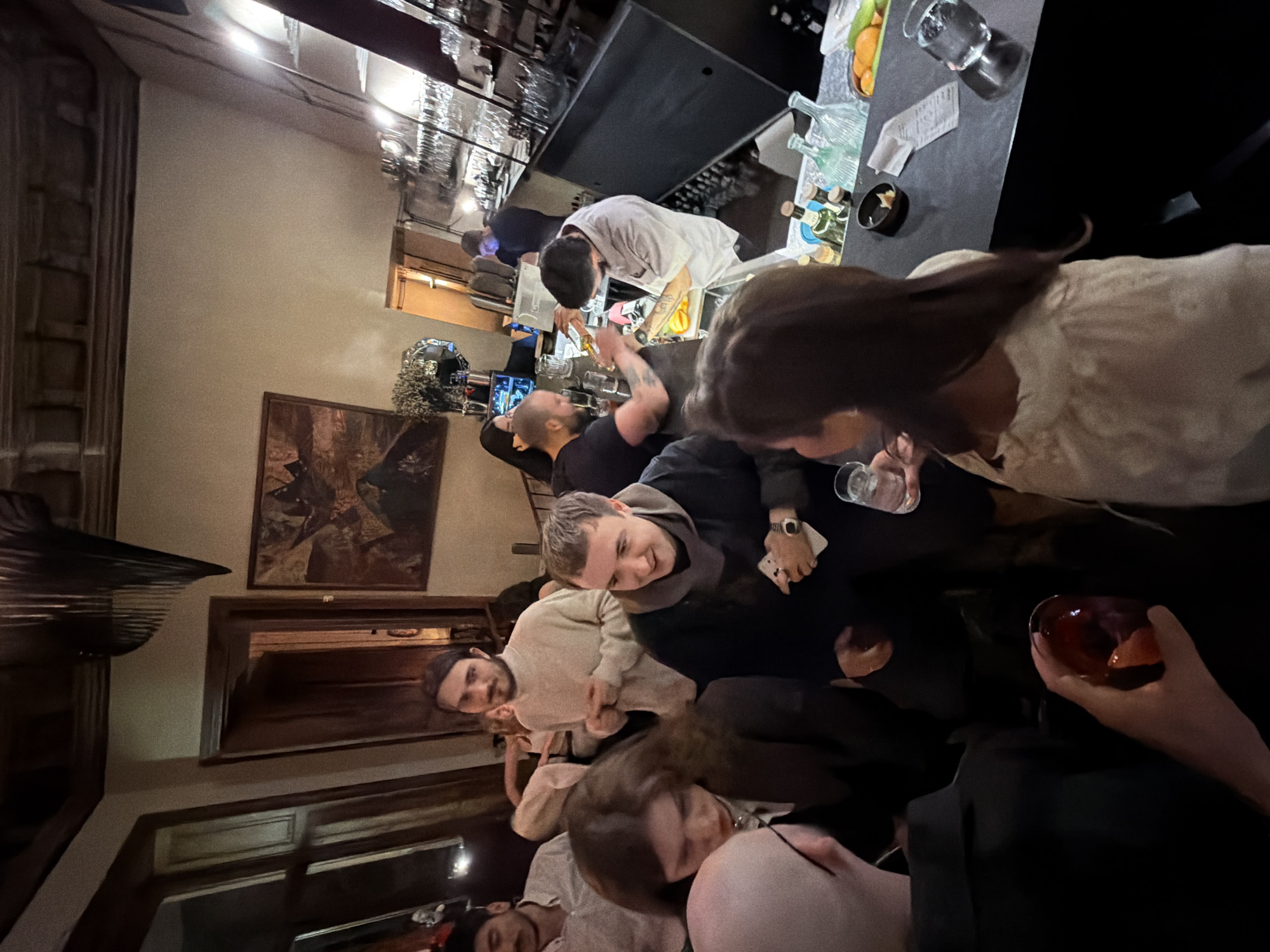 The image depicts a crowded indoor scene within Bouillon, a bar located in the city of Tbilisi, Georgia. The environment is dimly lit with warm artificial light and features exposed dark wooden ceiling beams, a dark modern hanging light fixture, and light-colored walls. A framed abstract or landscape painting with warm tones is visible on the far wall.
In the foreground, several individuals are partially visible. On the far left, a person with a bald head and glasses is seen from behind, facing right. To their right, a woman with long dark hair, wearing a light-colored, possibly embroidered, top, holds a clear glass with ice and a drink, looking towards the right. A hand in the lower center holds a glass with a dark red beverage and a visible citrus garnish.
The midground features numerous individuals standing and interacting. A man with dark hair and a beard, wearing a light cream-colored turtleneck sweater, stands centrally, looking forward. To his right, another man with short hair and a dark hooded top or scarf smiles, holding a white object (possibly a phone or card) in his left hand. Other patrons are visible further back, including a man with a mustache in a white shirt and a woman with long dark hair wearing a dark jacket.
In the background, the main bar area is active. A bartender, wearing a light-colored shirt or smock, stands behind a dark bar counter, actively preparing drinks with shakers and garnishes (visible oranges). Another person stands behind the bar to the left of the bartender. The bar counter displays bottles, shakers, glasses, citrus fruit segments, and a receipt. Behind the bar, extensive shelving holds numerous wine and cocktail glasses. Another individual in a dark shirt is partially visible further back, near an illuminated doorway.