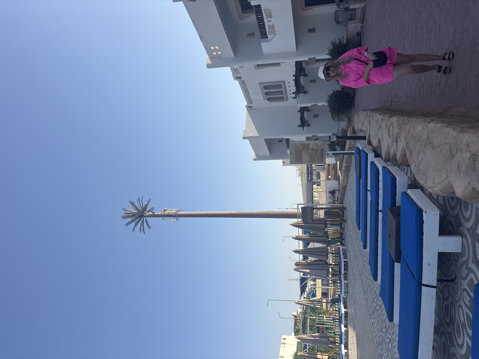 The image depicts a scene in Sharm El Sheikh, Egypt, on a bright, sunny day. In the foreground, a row of beach loungers with blue cushions are visible, lined up on a patterned tiled surface. Adjacent to the loungers, beige sun umbrellas are folded down.
To the left, a tall, cylindrical structure mimicking a palm tree stands prominently against the clear blue sky. Closer to the top, cellular antennas are discreetly attached to the faux palm foliage.
In the background, there is a multi-story white building with decorative elements. A woman wearing a bright pink outfit and a white hat stands on the sidewalk. She is carrying a black bag.