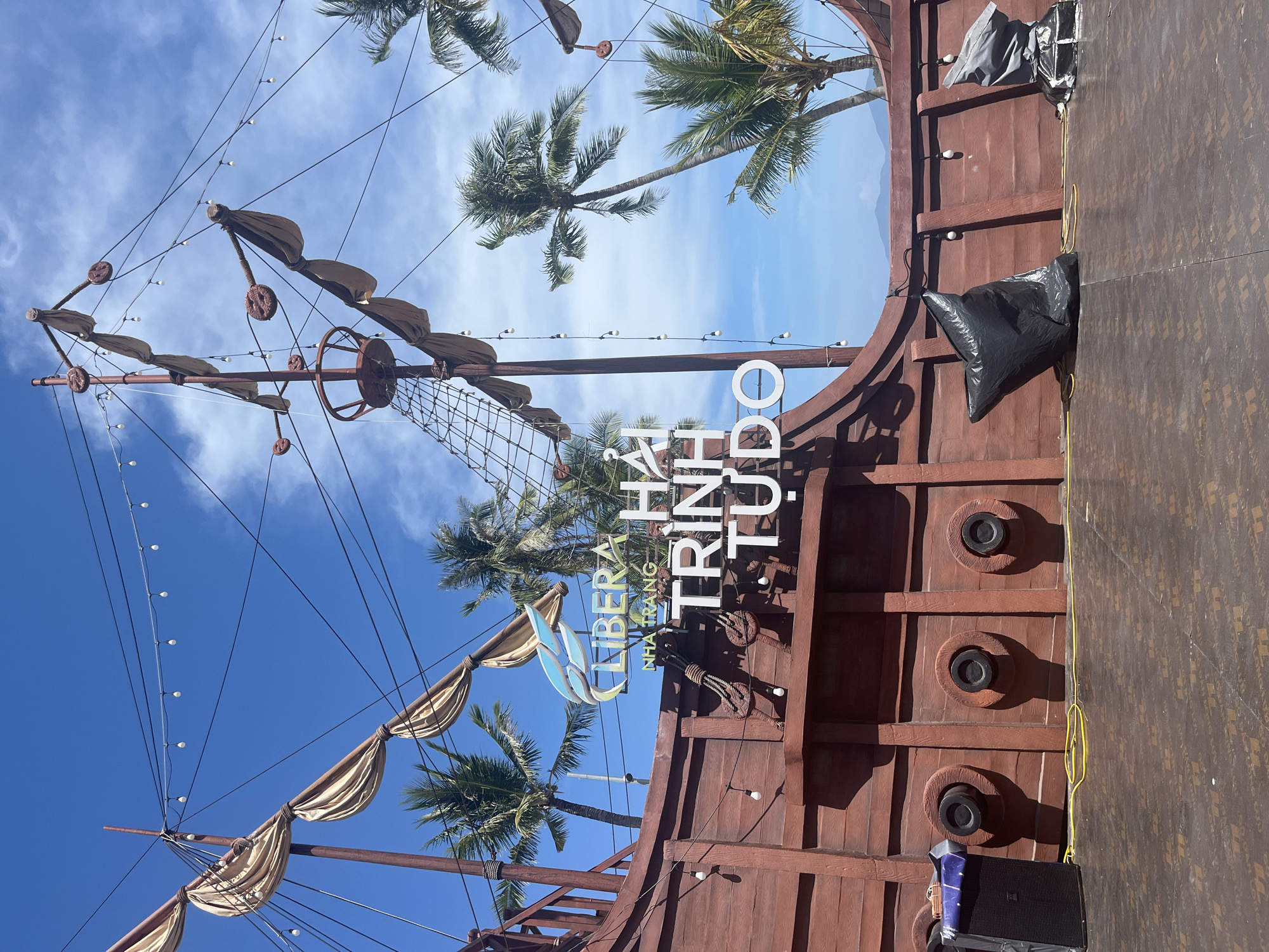 The image shows a section of a ship-themed structure in Nha Trang, Vietnam. The structure has a wooden façade with features resembling a ship's hull, complete with gunports. A mast with faux sails and rigging is visible. The signage reads "LIBERA NHA TRANG TRINH TUDO". Palm trees are visible in the background. There are also some bags and equipment on the right side of the image that could be related to event set up or construction. The sky is blue with scattered clouds.