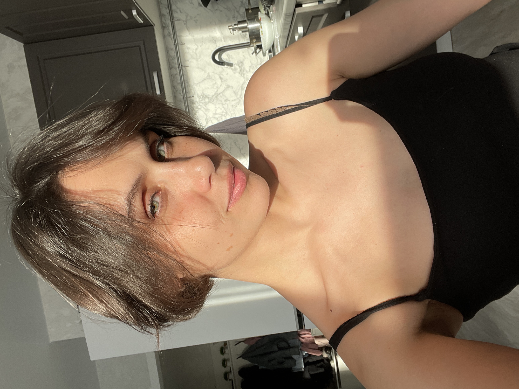 A young woman with short brown hair and green eyes is the primary subject of the image. She is wearing a black tank top with thin straps. The lighting suggests the image was taken during the day, likely indoors. The background features a kitchen setting with cabinets and a sink. It is not possible to determine if the image was taken in Izhevsk, Russia, or any other specific location.