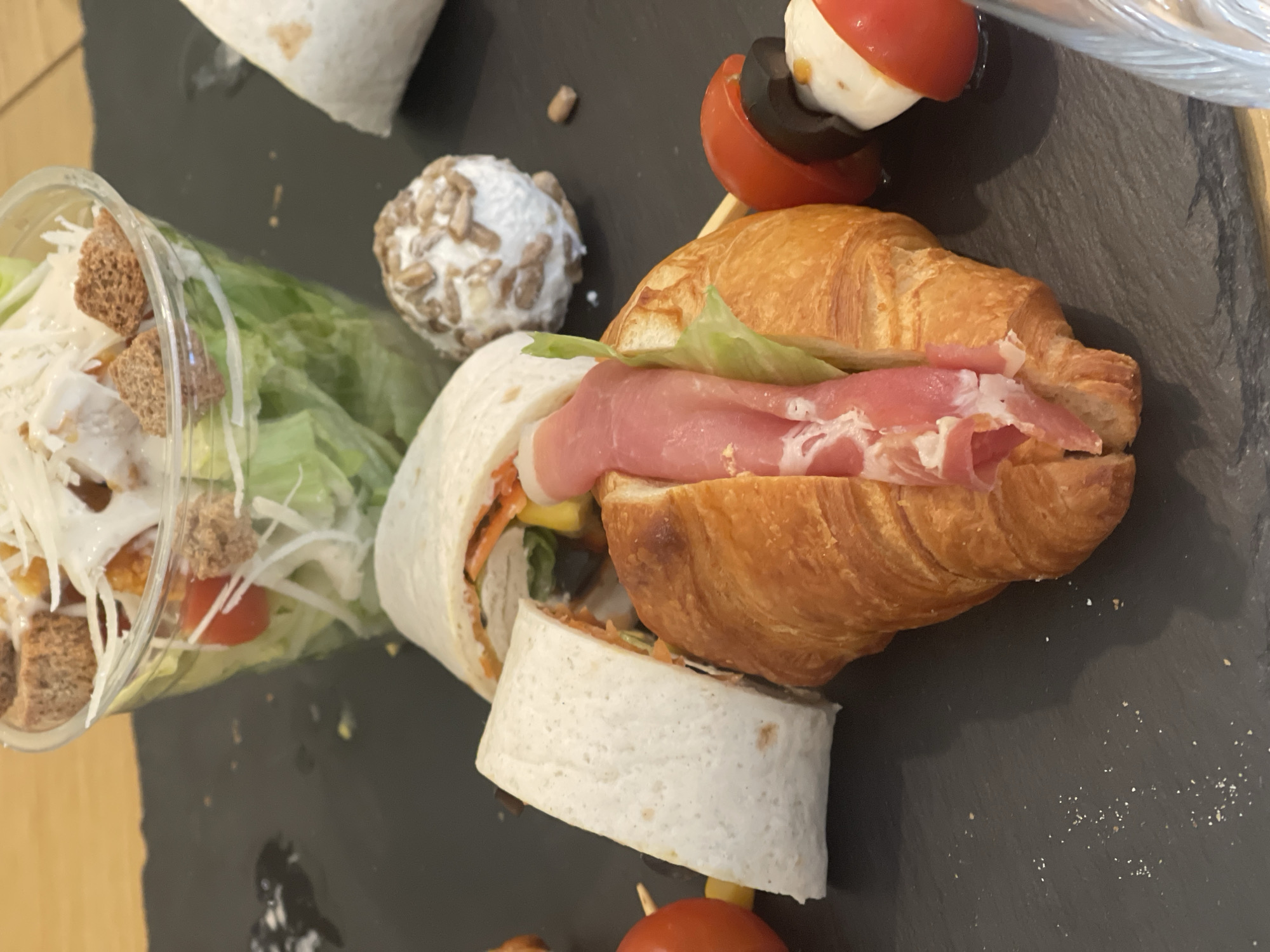 The image depicts a selection of food items arranged on a dark grey slate serving plate. The items include a salad in a clear plastic container with lettuce, shredded cheese, croutons, and what appears to be salad dressing. There is a wrap with a white tortilla cut into a piece and a croissant sandwich filled with what appears to be prosciutto and lettuce. There is a small ball covered with sunflower seeds and two skewers with tomato, mozzarella and black olive.
The location and city of Sofia, Bulgaria, cannot be determined from the image alone.