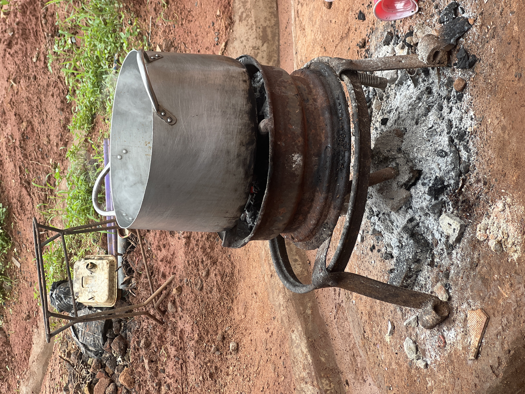 In Nkpor, Nigeria, a large aluminum pot is steaming as it sits atop a makeshift charcoal stove. The stove is constructed from a rusty metal rim, possibly from a car wheel, supported by a framework of bent metal rods. The stove is surrounded by gray ash and charred remnants of burnt wood. To the left of the stove, a metal rack holds a dirty white object, possibly a generator part, with a black bag and some rocks piled behind it. The ground is a reddish-brown dirt, with sparse green vegetation visible in the background. A red plastic cup lies on the ground to the right of the stove.