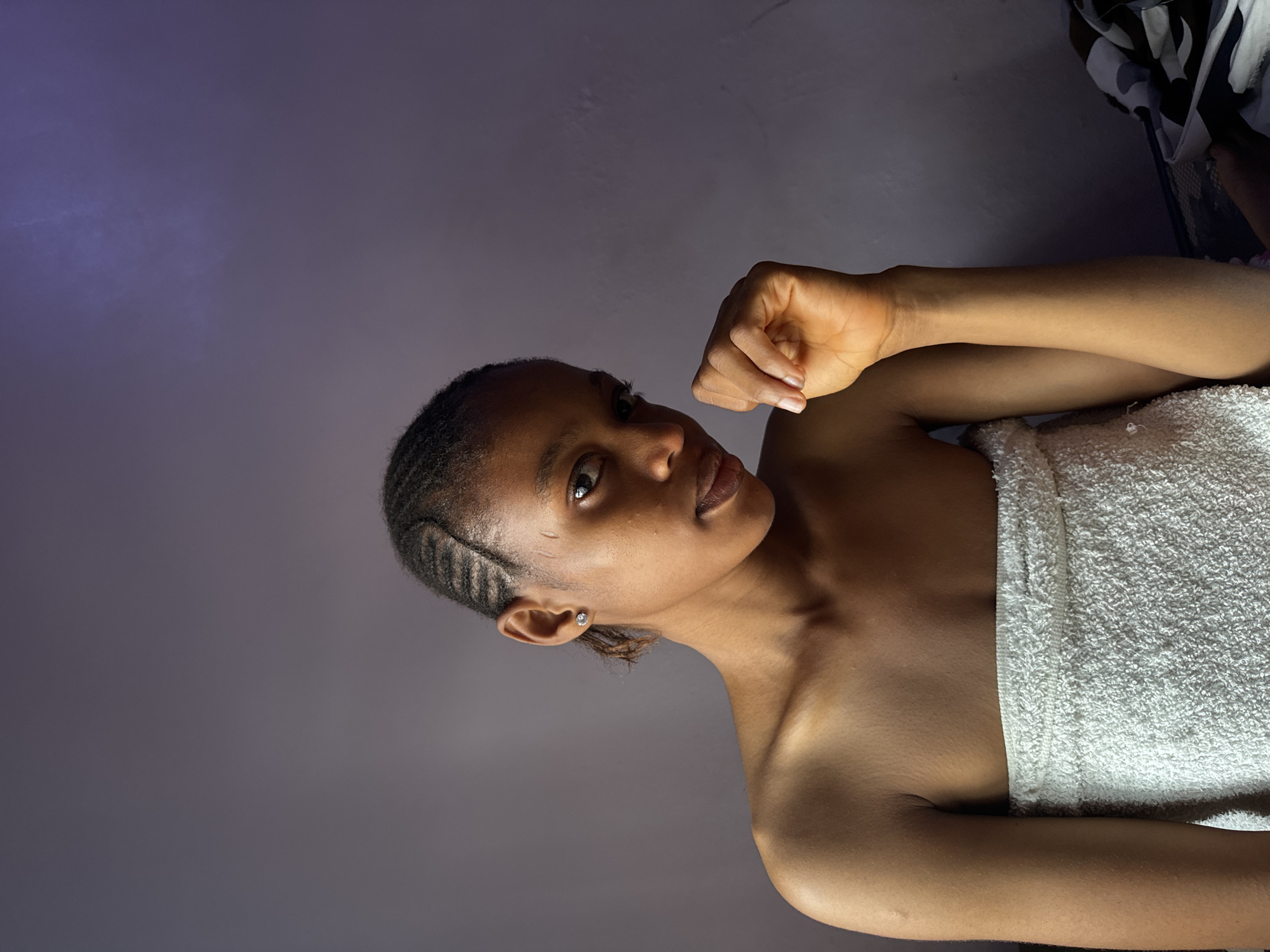 A young Black woman, with her hair styled in cornrows, is pictured from the chest up, wrapped in a white towel. She is looking directly at the camera with her head tilted slightly. Her right hand is raised and resting on her chin. The background is a plain, textured purple wall. The lighting suggests an indoor setting, possibly a bathroom or bedroom, with a soft light source illuminating her face from the front left and a faint purplish glow on the upper right side of the background. There is a hint of a patterned fabric on the right edge of the frame. The image does not contain any specific elements that would allow for definitive placement within Nkpor, Nigeria, or any other specific city.