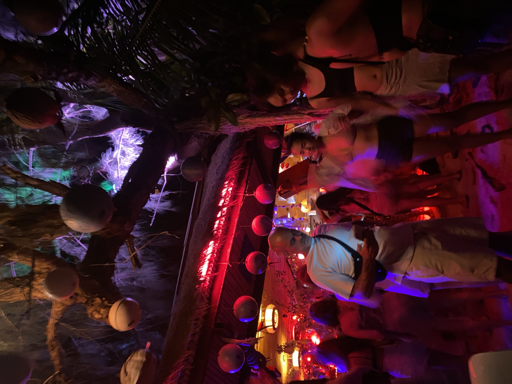 The image depicts a nighttime scene in Baan Sri Thanu, Thailand, possibly at a bar or restaurant with a tropical theme. Several people are visible, including a bald man in a white shirt and khaki shorts standing amongst the crowd. Other patrons are also present. The setting is decorated with spherical light fixtures resembling coconuts hanging from trees or structures. Red lights are also visible. The overall atmosphere appears to be lively and illuminated with colorful lighting.