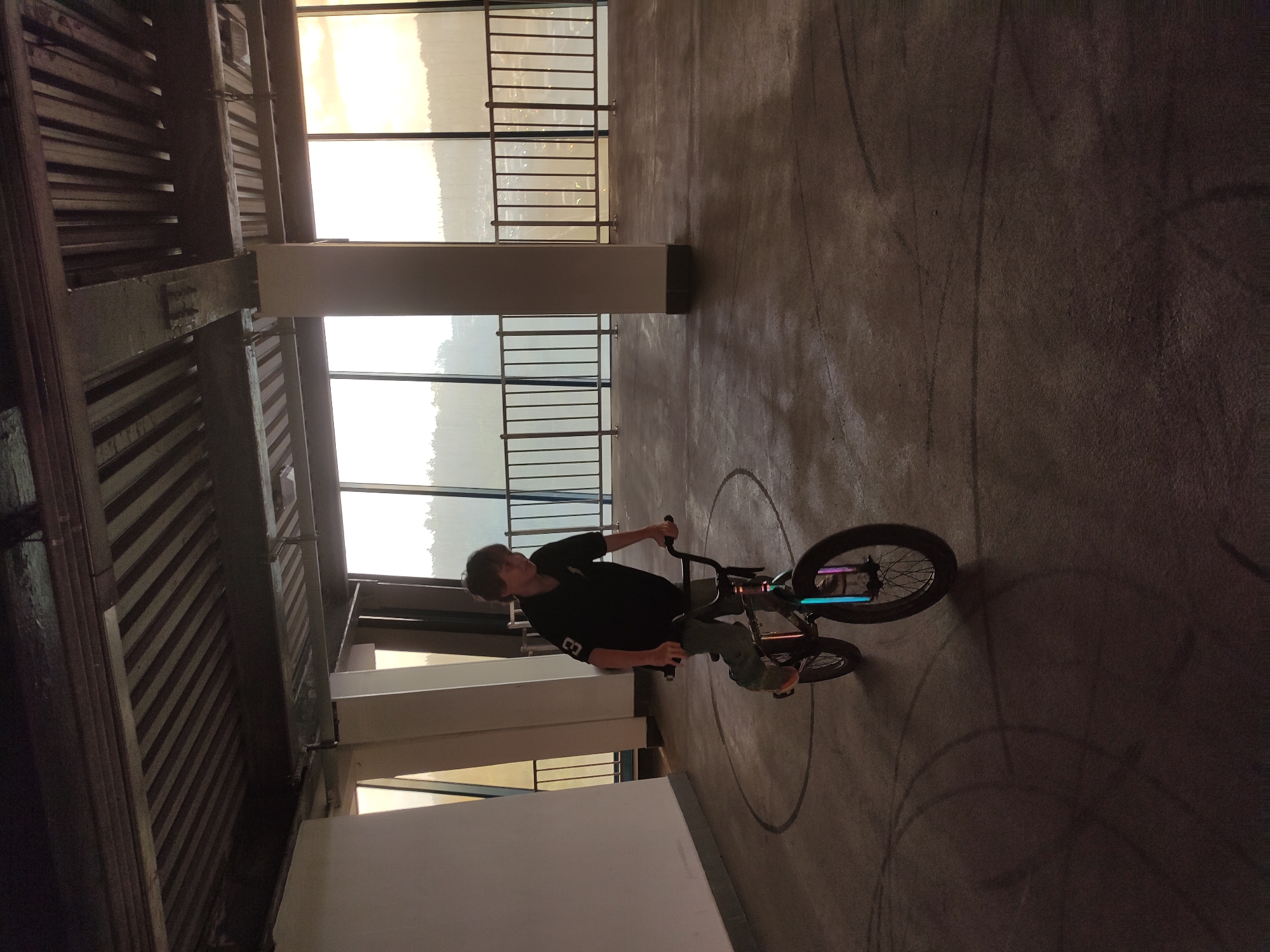 A rotated, medium indoor shot depicts a person on a BMX bike performing a trick in an urban setting. The person, wearing a dark t-shirt, light-colored pants, and shoes, is positioned on the bike within a circular marker on the concrete floor. To the left, a large structure with visible architectural elements including support beams, window panes, and metal guardrails is observed. Beyond the windows, a bright outdoor environment is visible, potentially indicating daylight conditions. The general atmosphere suggests an enclosed, perhaps multi-story parking area or similar space.