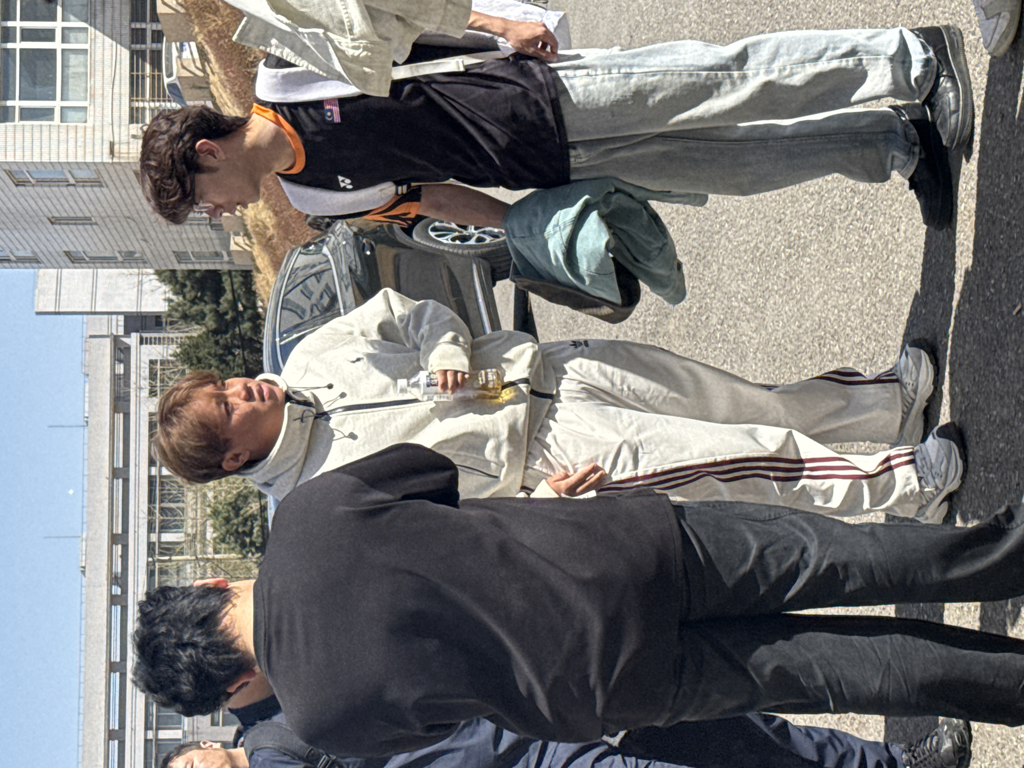In Zhongtun, China, a group of people stands on an asphalt surface, likely a street or parking area. One individual wears a black shirt with orange trim and light-wash jeans. Another is dressed in a light-colored tracksuit with maroon stripes. A third person wears black pants and a black top. At least one more individual is partially visible, wearing a blue jacket. A car is partially visible in the background. Some of the people appear to be holding or interacting with an item or object. The environment includes building facades, trees, and a blue sky.