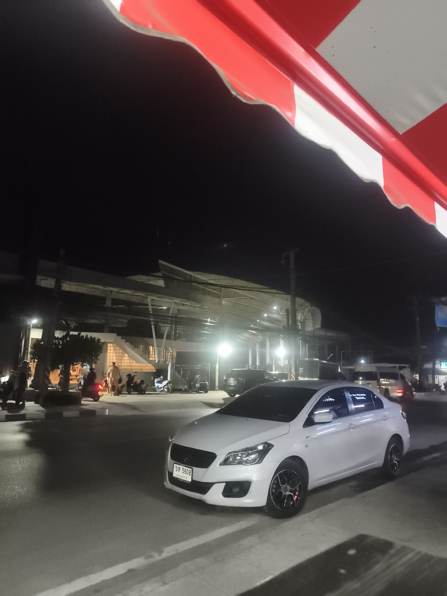 A white sedan is parked on a street in Ko Samui District, Thailand at night. The car has a license plate with Thai characters and the number 5608. In the background, there are several motorcycles parked, some people walking, and a building with lights on. A red and white striped awning is visible in the upper portion of the image. The street appears to be paved and there are streetlights illuminating the scene.