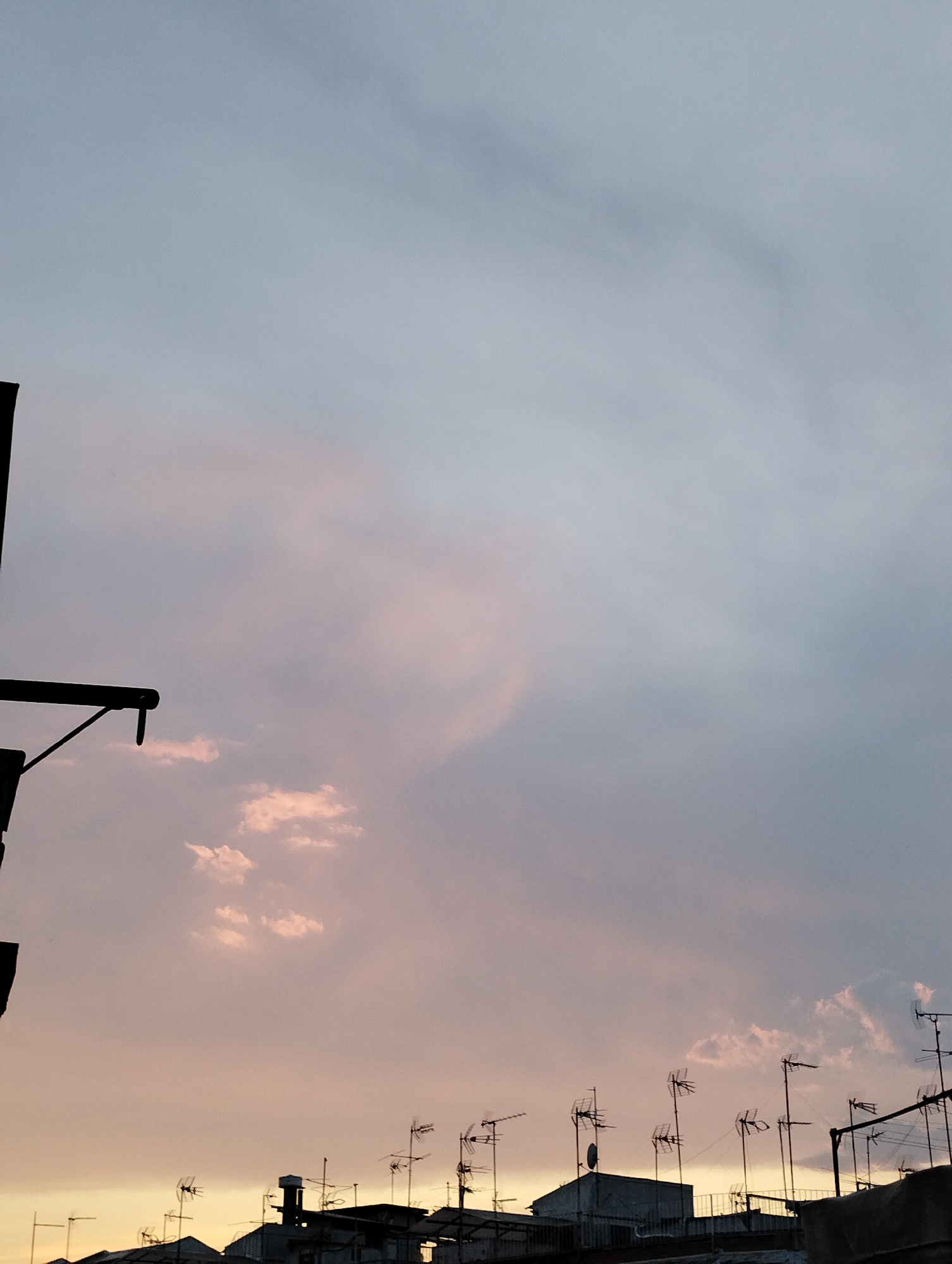 The image depicts an urban scene at dusk in Unknown, Unknown. The sky is a mix of pale blue and pink hues, suggesting a sunset or sunrise. Clouds are scattered throughout, some tinged with pink. The foreground shows rooftops densely populated with television antennas. A dark, silhouetted structure is visible on the left side of the frame.