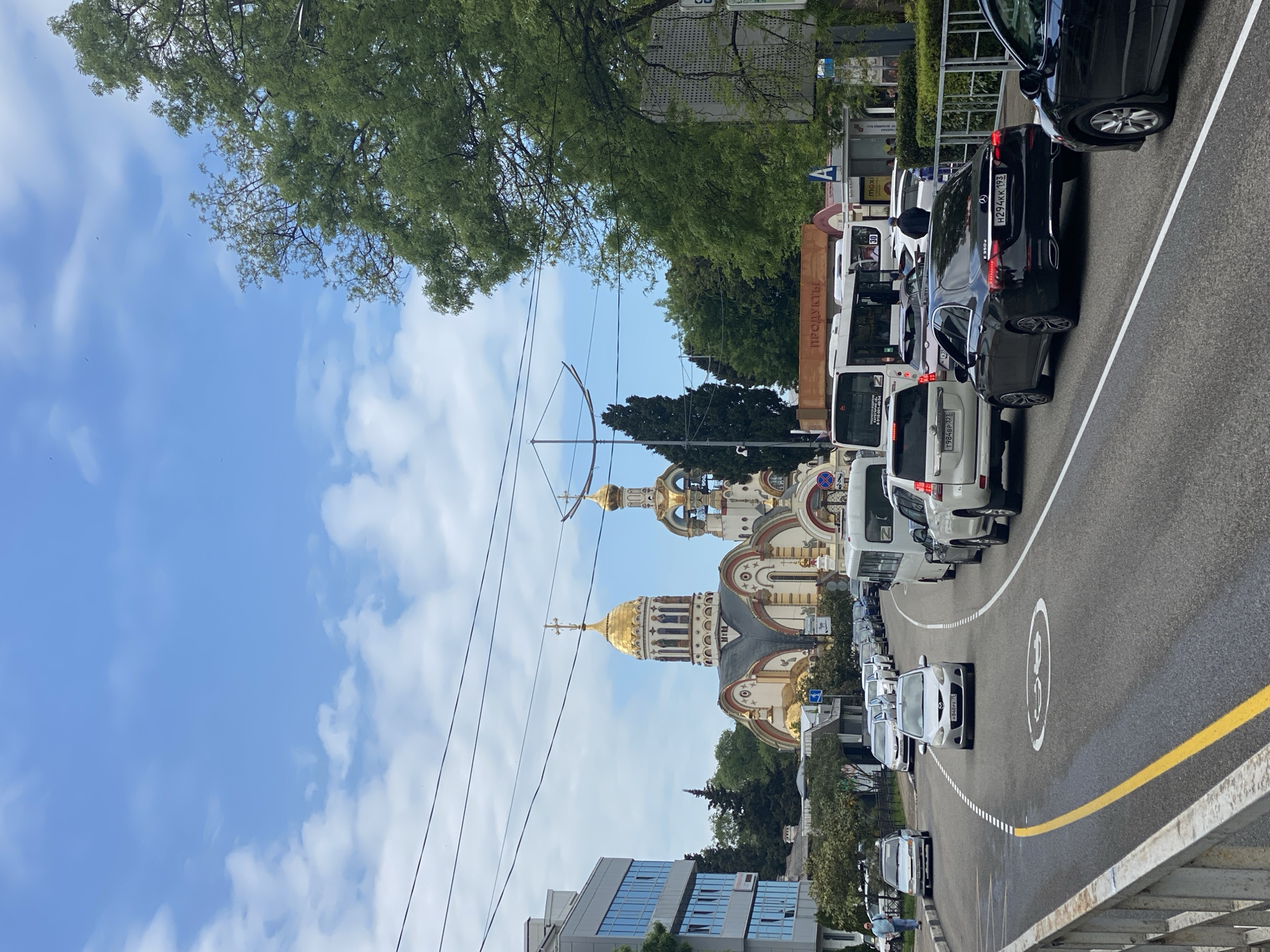 The image depicts a street scene in Sochi, Russia, featuring the Cathedral of Michael the Archangel in the background. The cathedral, characterized by its golden domes and ornate architecture, stands prominently behind a line of parked and moving vehicles on the street. The street is marked with a yellow line, a dashed white line, and a painted speed limit circle. Above, power lines stretch across the sky, which is partly cloudy. Trees and buildings are visible along the sides of the road.