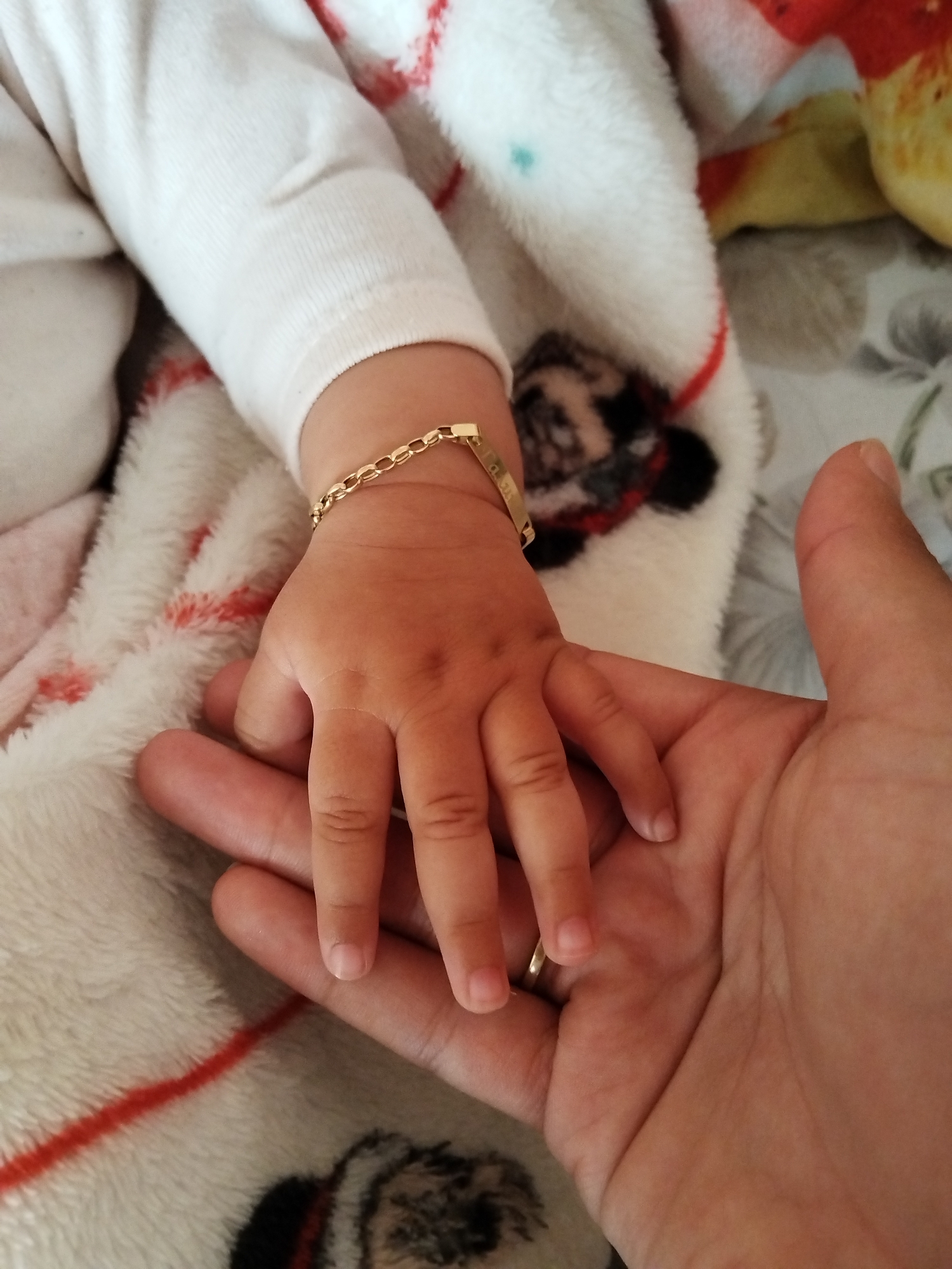 A close-up shot displays a baby's hand, adorned with a gold bracelet, resting gently on an adult's hand. The baby wears a white, long-sleeved garment and lies on a soft, patterned blanket with hints of red and black. The background remains out of focus. No location is discernible.