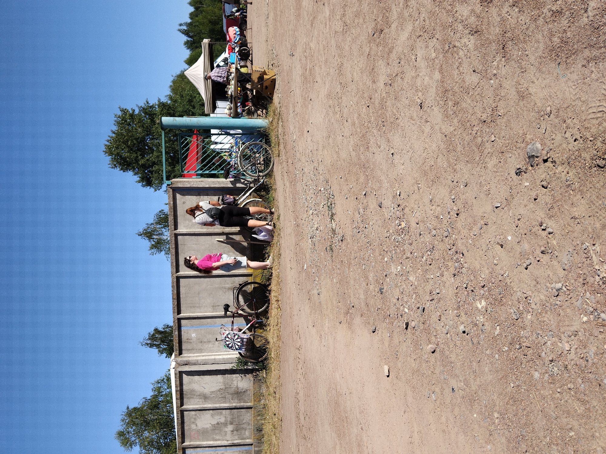 The image depicts an outdoor scene, likely a flea market or an informal gathering, under a clear blue sky. In the foreground, a dirt and gravel path fills the lower half of the frame. Behind this path, a concrete wall stretches horizontally across the image.
Along the wall, several bicycles are visible. One bicycle, on the left, has a dartboard attached to its spokes, with colorful points. Two women are standing near bicycles. The woman on the left wears a bright pink t-shirt and light-colored shorts. The woman on the right wears a striped t-shirt and dark shorts, and has a backpack. Further to the right, a green metal gate stands open, revealing a glimpse of a red canopy behind it.
Beyond the gate and the concrete wall, a market stall is set up under a white tent. A person, appearing to be a vendor, is behind the stall, interacting with a customer. Various items are displayed on tables at the stall, suggesting goods for sale. Several vehicles, including vans, are parked in the background, and a dense line of trees forms the horizon. The overall environment suggests a sunny day at a public gathering space. The scene is located in the city of Irshansk, Ukraine.