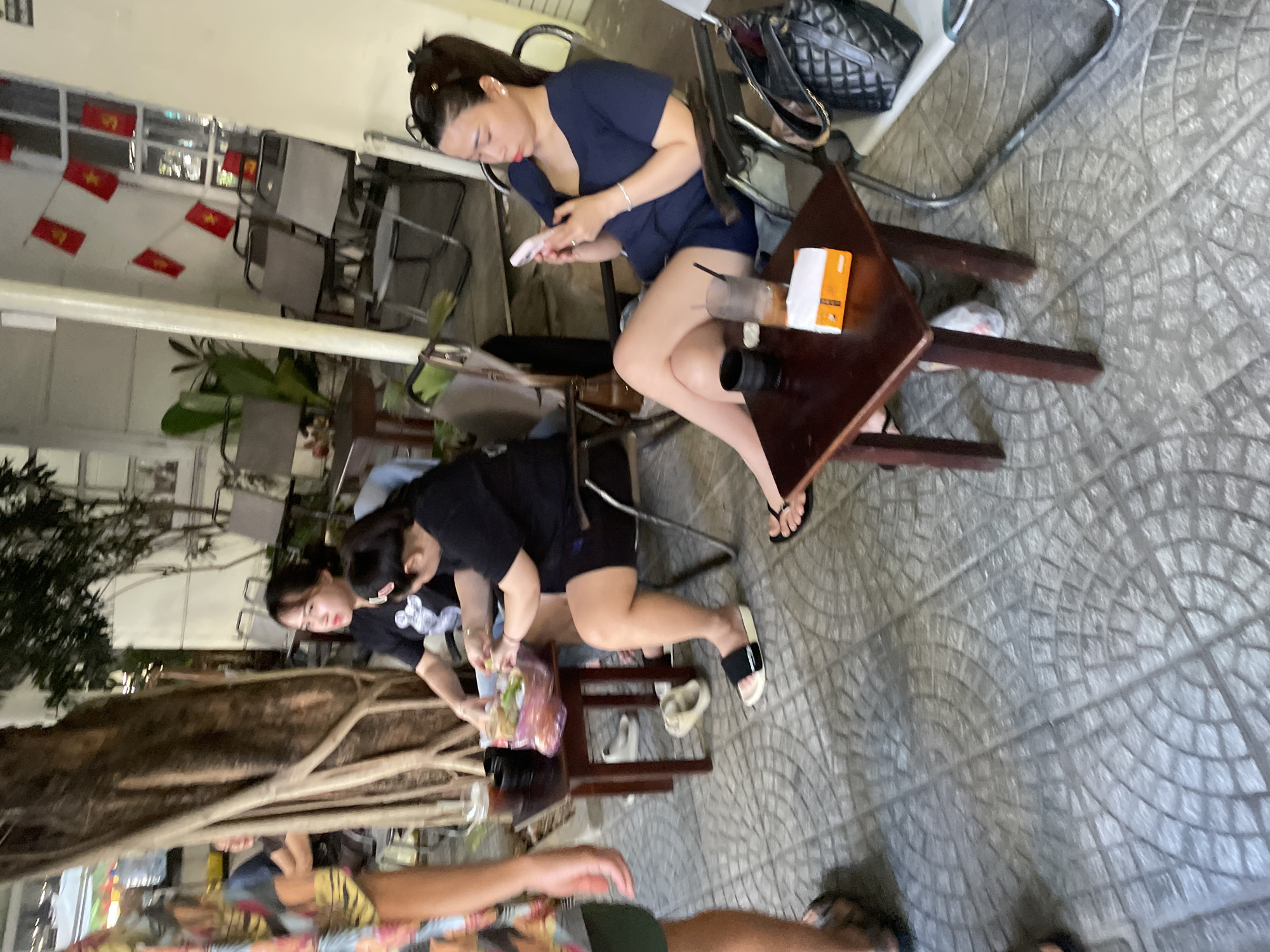 The image depicts an outdoor seating area, likely a cafe or restaurant, in Đà Nẵng, Vietnam, with a patterned concrete floor. Three women are seated at tables. On the left, two women in black tops are seated at a dark wooden table. One is looking towards the camera, and the other is looking down at something on the table. A bag of food and a drink are on their table. In the center-right, a woman in a dark blue top is seated at another dark wooden table, looking at her phone. A drink and a box of tissues are on her table. A black quilted handbag is on the chair next to her. Chairs and tables are visible in the background, along with plants and Vietnamese flags displayed on a window. A large tree trunk is on the left side of the image.