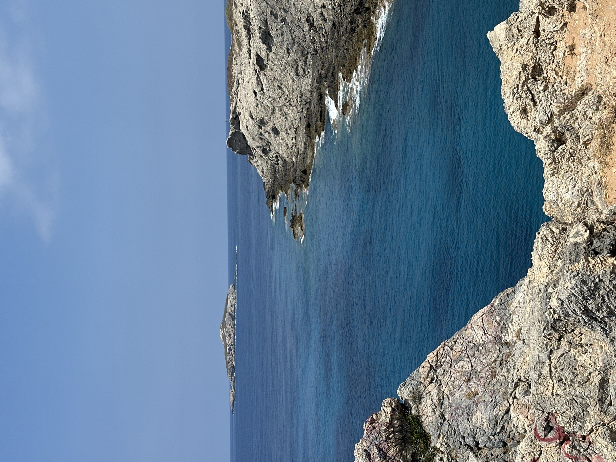The image captures a coastal scene with rugged, rocky terrain meeting the deep blue sea. In the foreground, large, weathered rocks with visible cracks and some red spray paint mark the edge of a cliff. The rocks are a pale, sandy color with gray and pinkish hues. Beyond the immediate foreground, the sea stretches out, its color transitioning from a vibrant turquoise near the shore to a deeper indigo further out.
A prominent feature in the middle ground is a rocky coastline that curves into the sea, with waves gently breaking against its base. Further in the distance, a small, elongated island with rocky outcrops and sparse vegetation sits on the horizon under a clear, pale blue sky. The overall impression is one of a remote and natural coastal landscape. The location is identified as being in the vicinity of Rizokarpaso, Northern Cyprus. No people or specific structures are visible in the image.