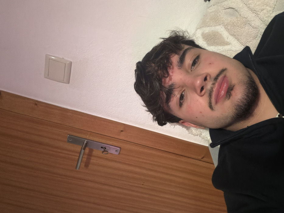 The image portrays a young male in an indoor environment in Vila Real, Portugal. The male, positioned in the lower right, has dark, wavy hair, a reddish mark on his forehead, light brown eyes, a mustache, and a short goatee. He is wearing a black collared, zippered top and is observed in a reclining posture, with his head resting on a light-colored, textured object, likely a pillow or blanket. He is looking directly towards the viewer. The background on the left side features a light brown wooden door with a visible grain, fitted with a metallic door handle and a key inserted into its keyhole. To the right of the door and positioned above the male's head, a textured, off-white wall is present, on which a light beige square electrical switch or outlet cover is mounted.