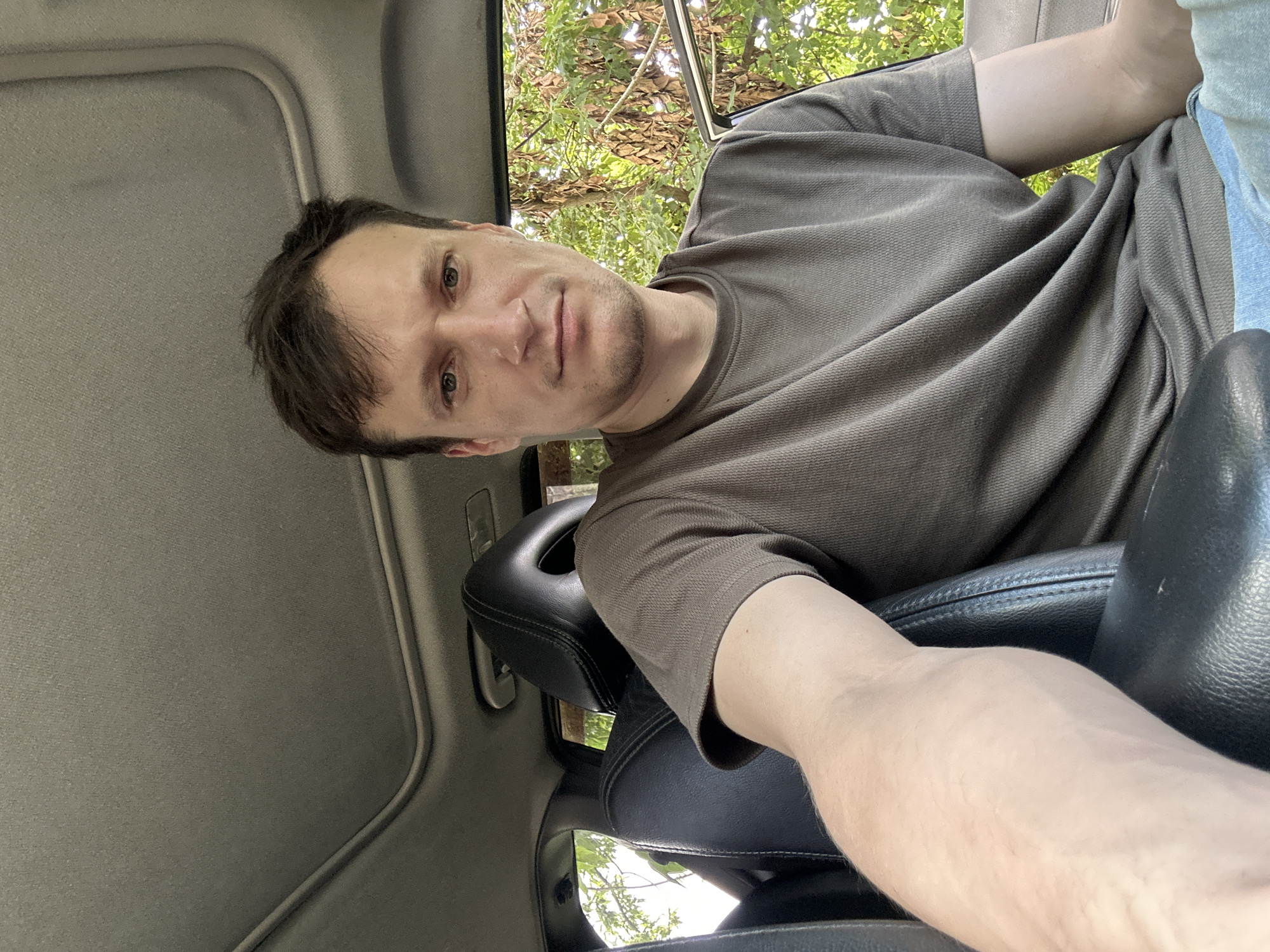 A man with brown hair and green eyes is sitting in the driver's seat of a car. He is wearing a brown t-shirt and blue jeans. The car has black leather seats and a sunroof. The background shows trees and greenery, suggesting the car is parked outdoors. The image is taken from a slightly high angle, looking down at the man. The location is Makiv, Ukraine.