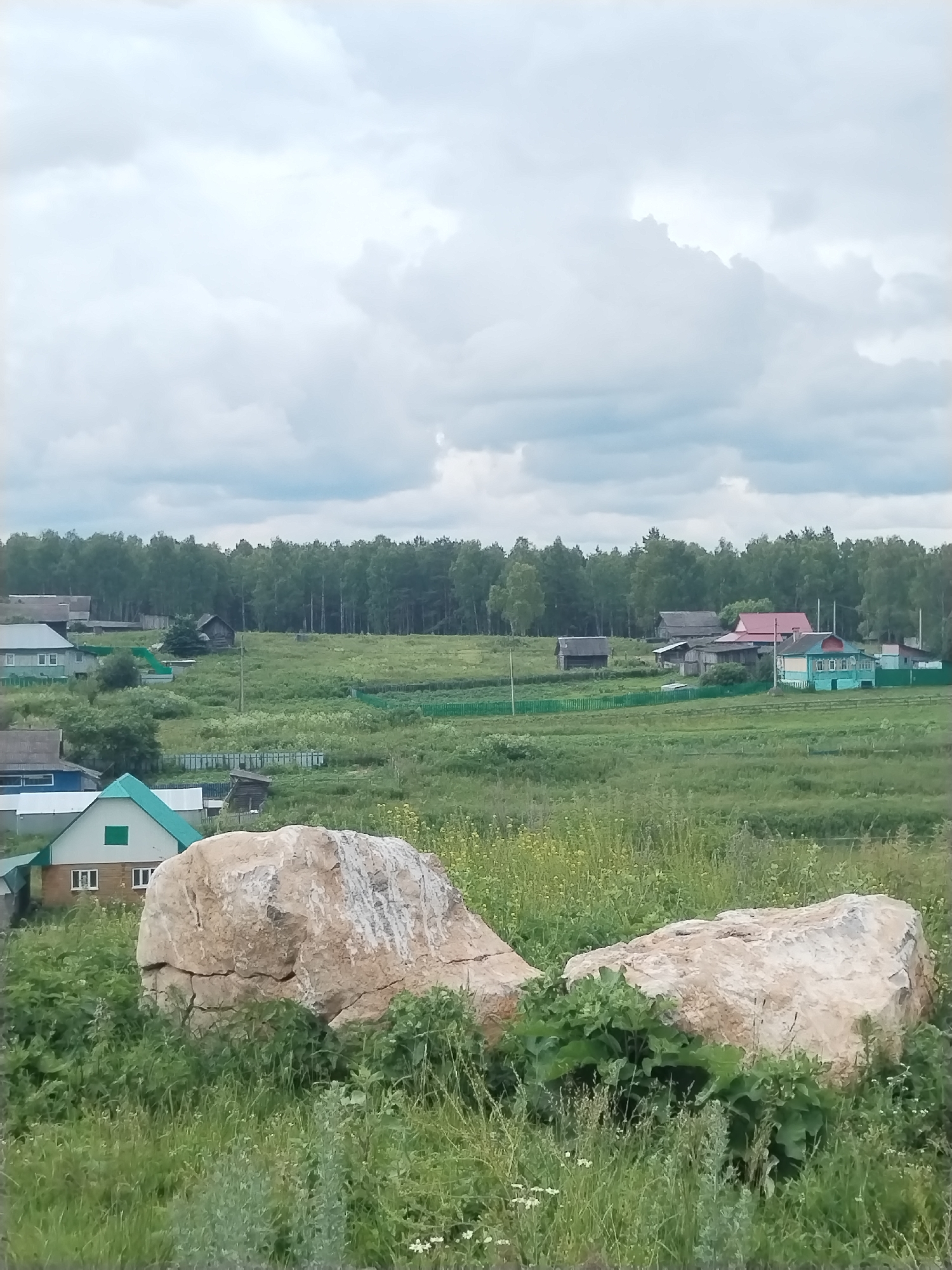The image shows a rural landscape with a cloudy sky. In the foreground, two large, light-colored boulders sit on a grassy slope. Behind the boulders, a village with several houses and outbuildings is visible. The houses are a mix of styles, with some having green roofs and others painted in various colors. A dense forest borders the village in the background. The overall impression is one of a peaceful, rural setting in the village of Idrisovo, Russia.