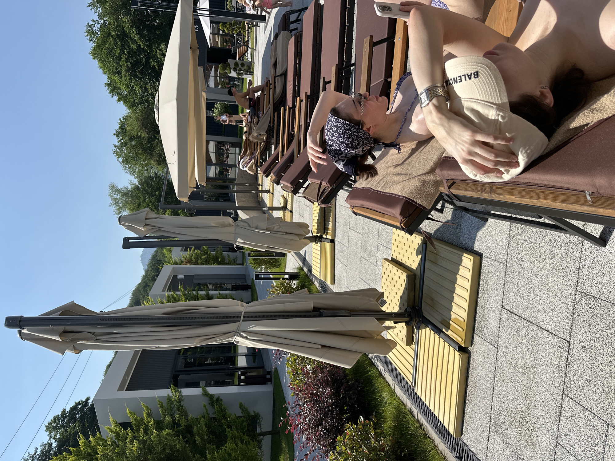 The image shows a resort area in Verkhnyaya Saniba, Russia. In the foreground, two women are lounging on recliners under a clear blue sky. The woman closest to the camera is wearing a bikini and a bandana, holding a smartphone. The woman behind her is wearing a baseball cap. Several closed umbrellas are positioned around the recliners. In the background, there are modern buildings with large windows, lush green trees, and a mountain range. The overall scene depicts a relaxing day at an outdoor resort.