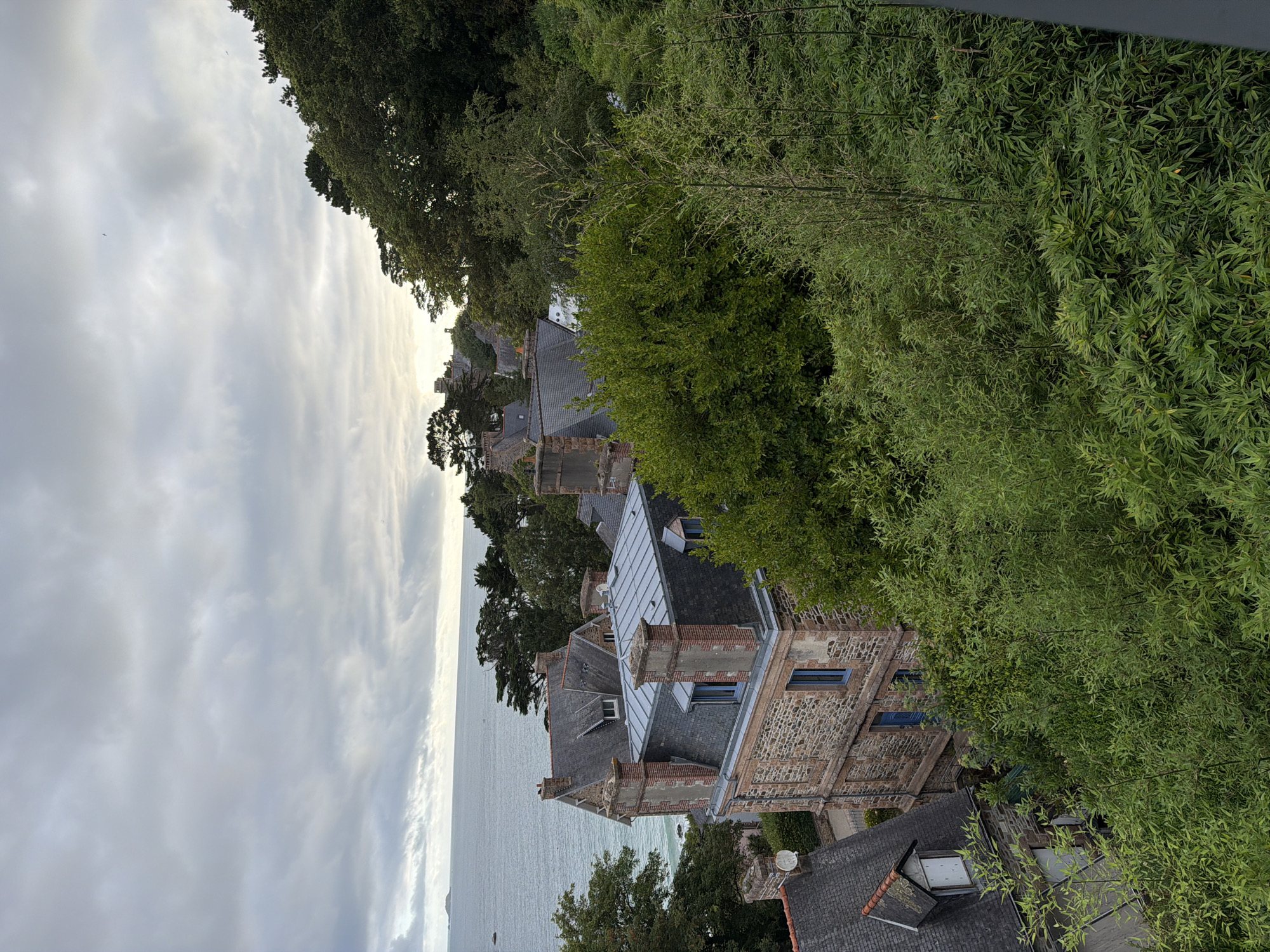 The image is an elevated, eye-level exterior shot of the coastal town of La Clarté, France, under a cloudy sky. The scene features several stone and slate-roofed buildings with red brick accents, nestled amongst lush green trees and vegetation. The ocean is visible in the background, with a small island in the distance. The architecture suggests a historical European coastal village. There are no people or animals present.